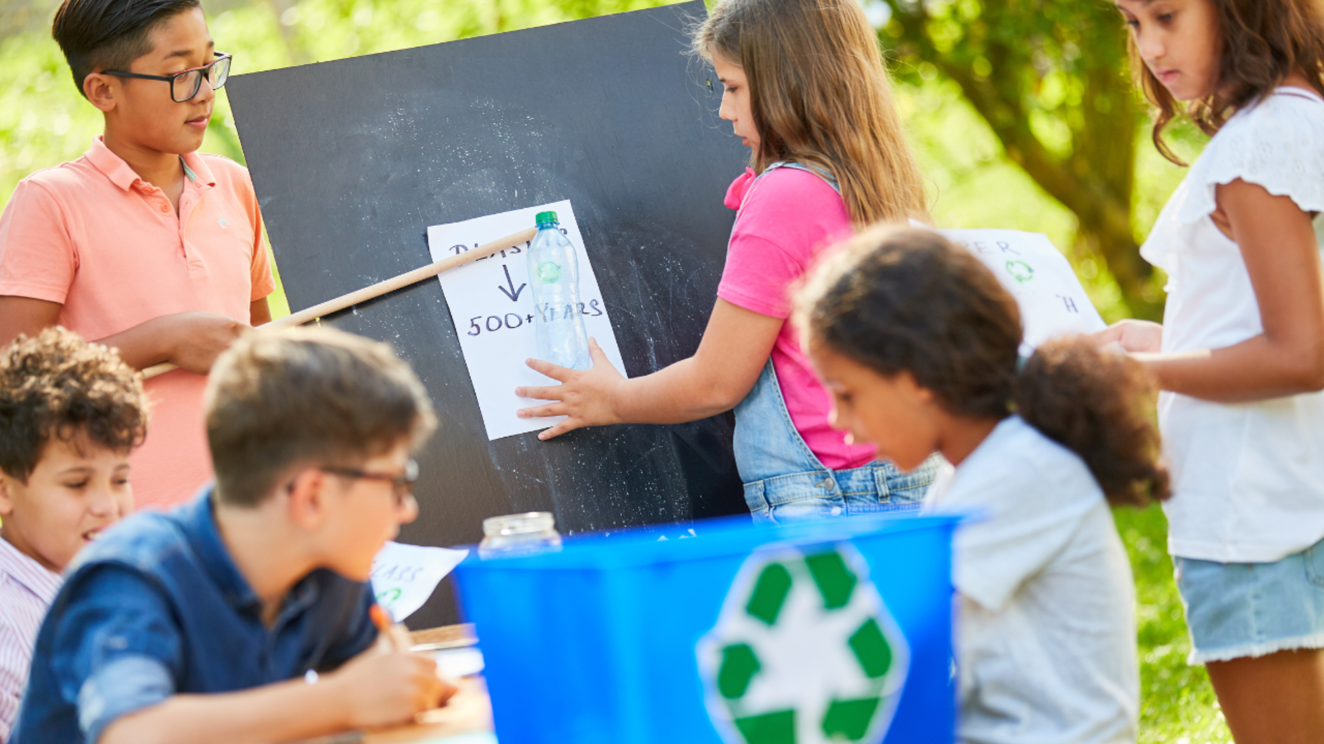 Des enfants sont rassemblés en plein air autour d'un tableau noir et d'une poubelle de recyclage bleue. Une fillette colle au tableau une feuille sur laquelle est dessinée une bouteille, tandis qu'un autre enfant la montre du doigt. D'autres sont assis, en train d'écrire ou de travailler sur des projets.