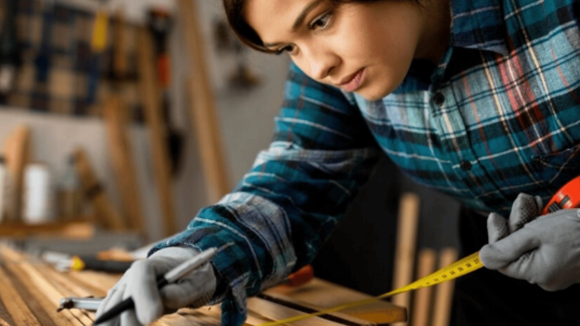 Une personne portant une chemise à carreaux et des gants mesure une pièce de bois à l'aide d'un mètre ruban, tout en se concentrant sur son travail dans un atelier de menuiserie.