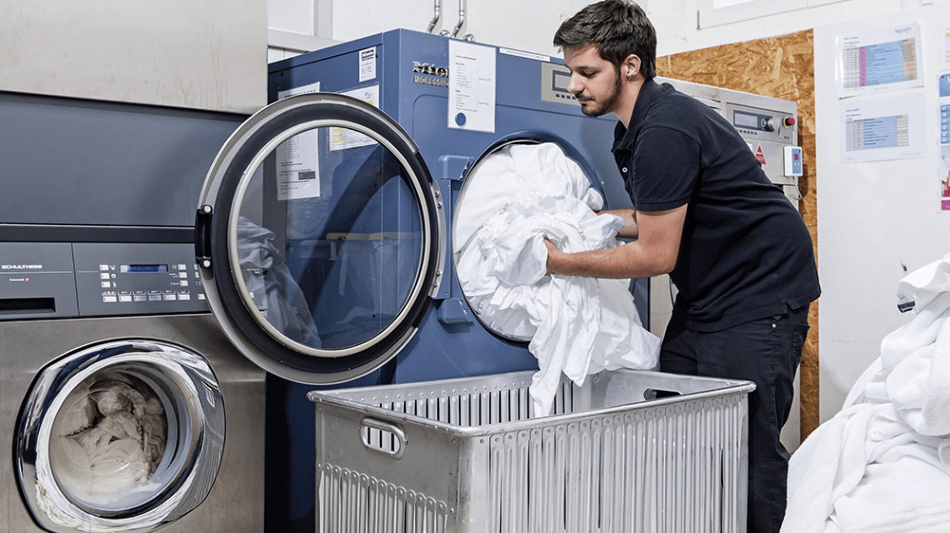 Un homme charge du linge blanc dans une grande machine à laver à chargement frontal dans une blanchisserie, avec un chariot métallique rempli de linge devant lui. Une autre machine à laver se trouve à gauche.