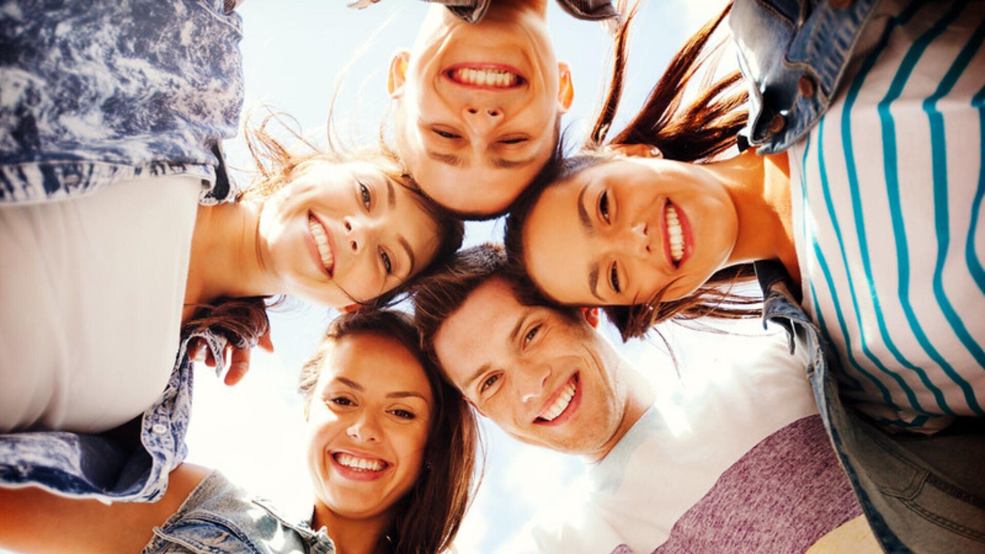 Cinq jeunes adultes se tiennent en cercle et sourient à l'appareil photo, la tête rapprochée, avec en arrière-plan un ciel d'un bleu éclatant.