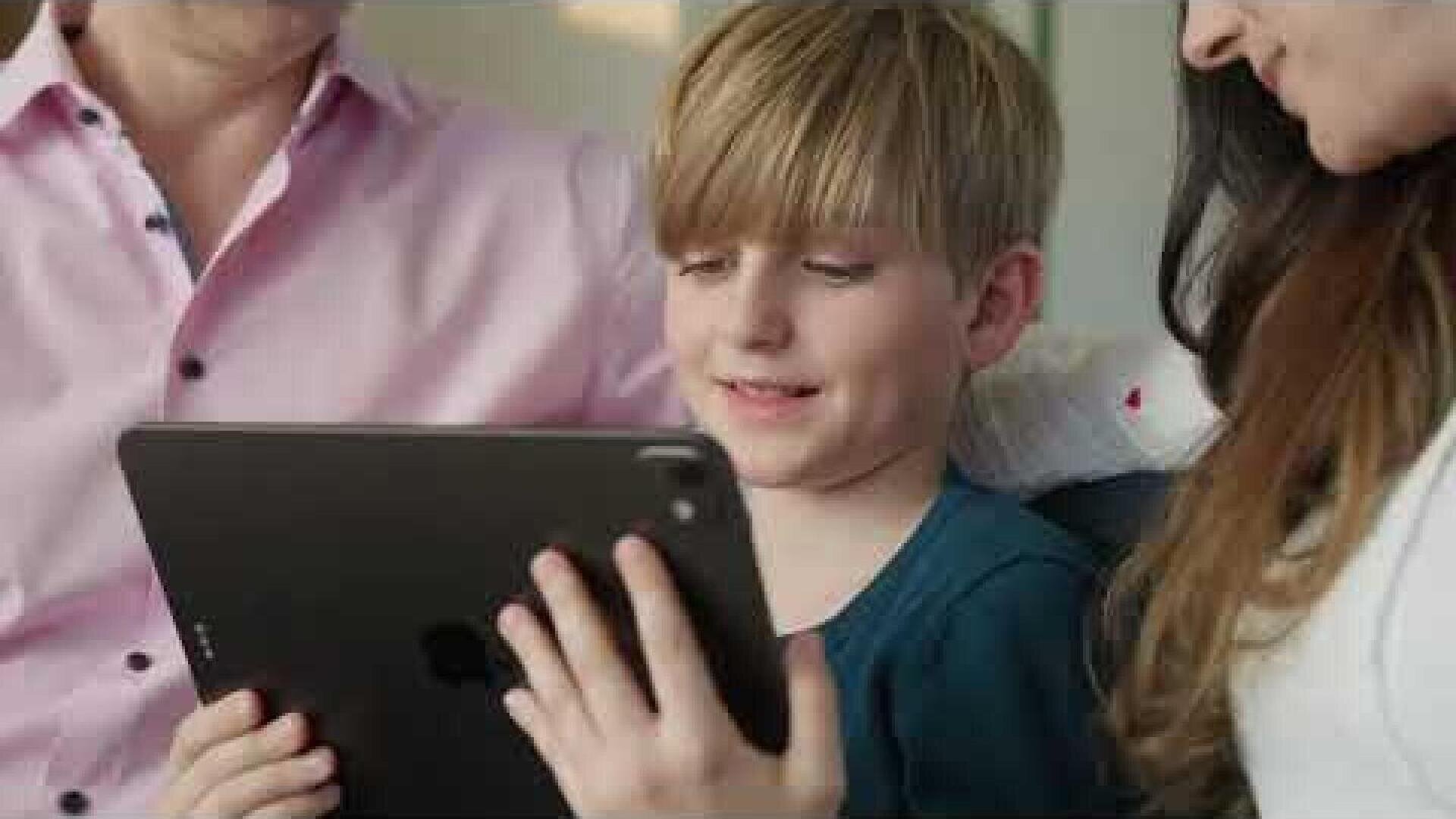 Un jeune garçon sourit, une tablette à la main, assis entre deux adultes. Le trio semble apprécier ensemble ce qui s'affiche à l'écran, dans un cadre lumineux et confortable.