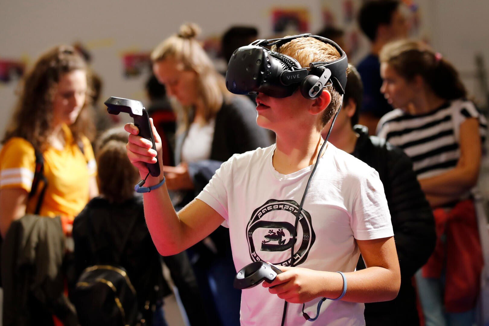 Un garçon portant un casque de réalité virtuelle et tenant des manettes explore une expérience de RV dans une salle bondée avec d'autres personnes en arrière-plan.