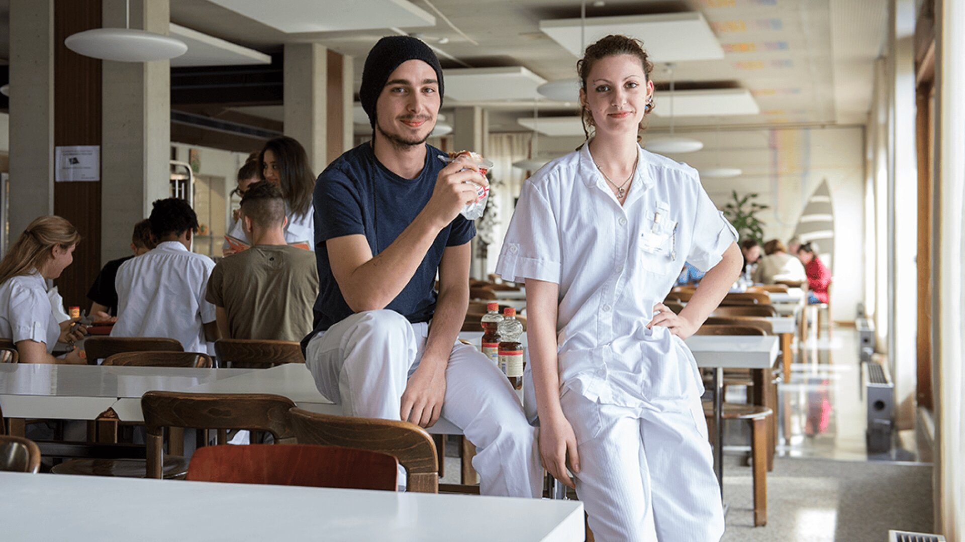Deux jeunes adultes en uniforme blanc posent et sourient dans une cafétéria lumineuse. L'un est assis sur une table et tient un sandwich, l'autre est debout à côté de lui. D'autres personnes sont assises et discutent à l'arrière-plan.