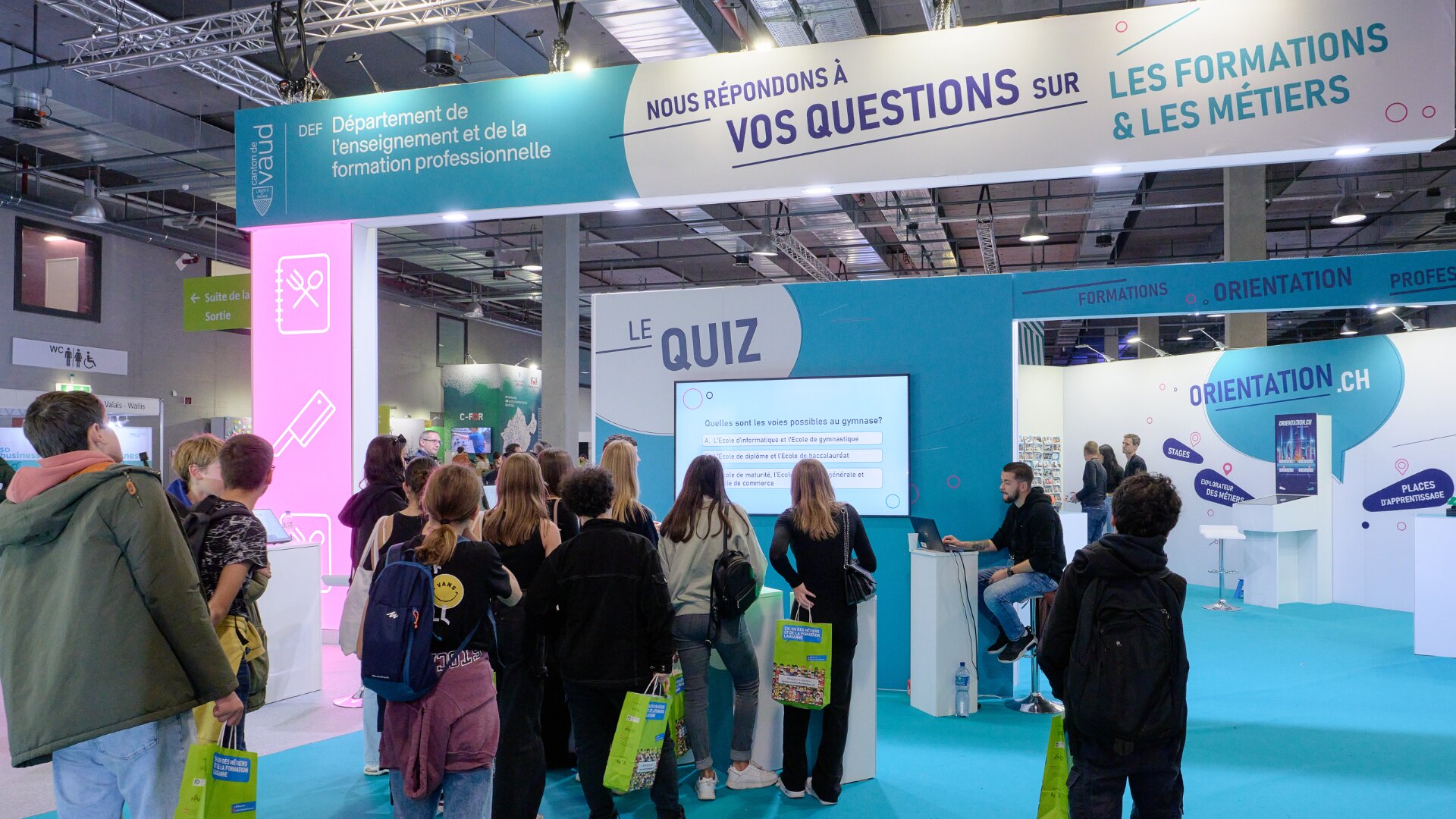 Un groupe de personnes se tient devant un écran de quiz lors d'un salon de l'éducation, où des stands proposent des informations sur les formations et les carrières. Des bannières et des panneaux sur la formation professionnelle sont visibles au-dessus et autour du stand.
