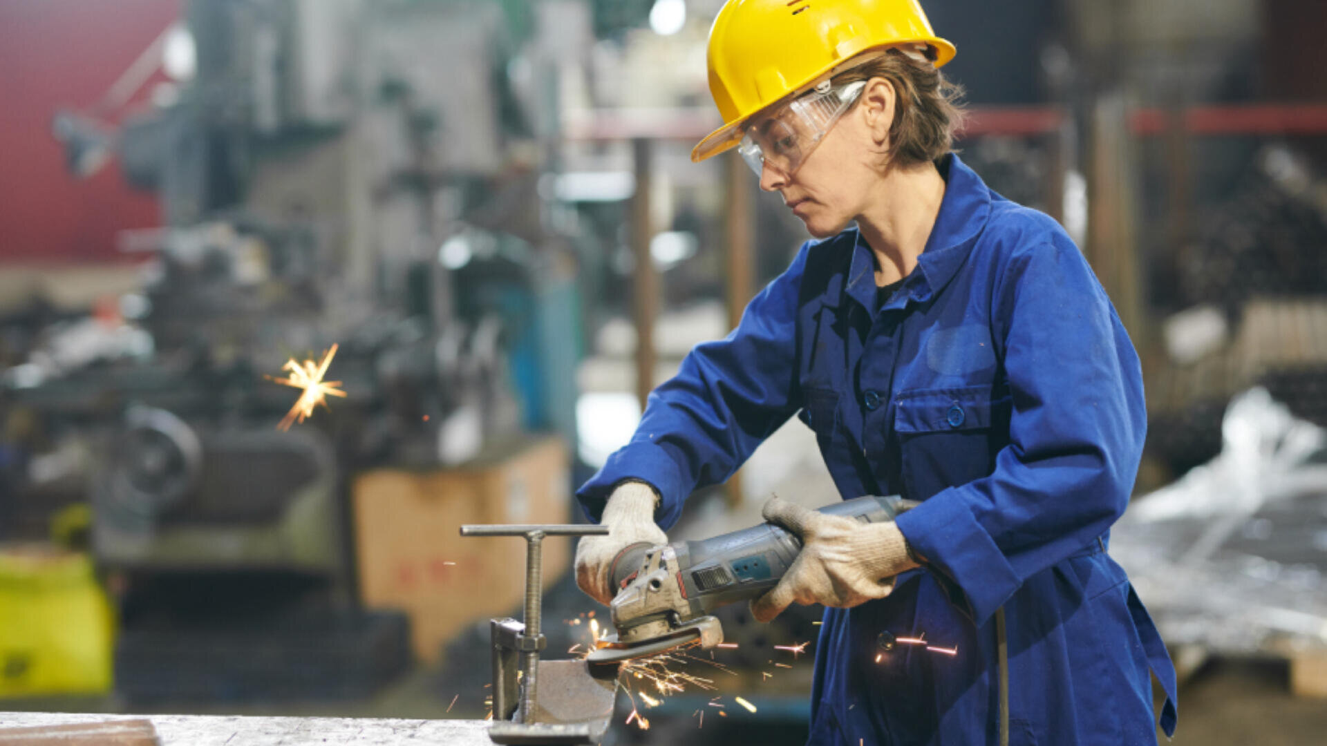 Une personne portant un casque jaune, des lunettes de sécurité, des gants et une combinaison bleue utilise une meuleuse d'angle sur un objet métallique, avec des étincelles dans un atelier industriel.
