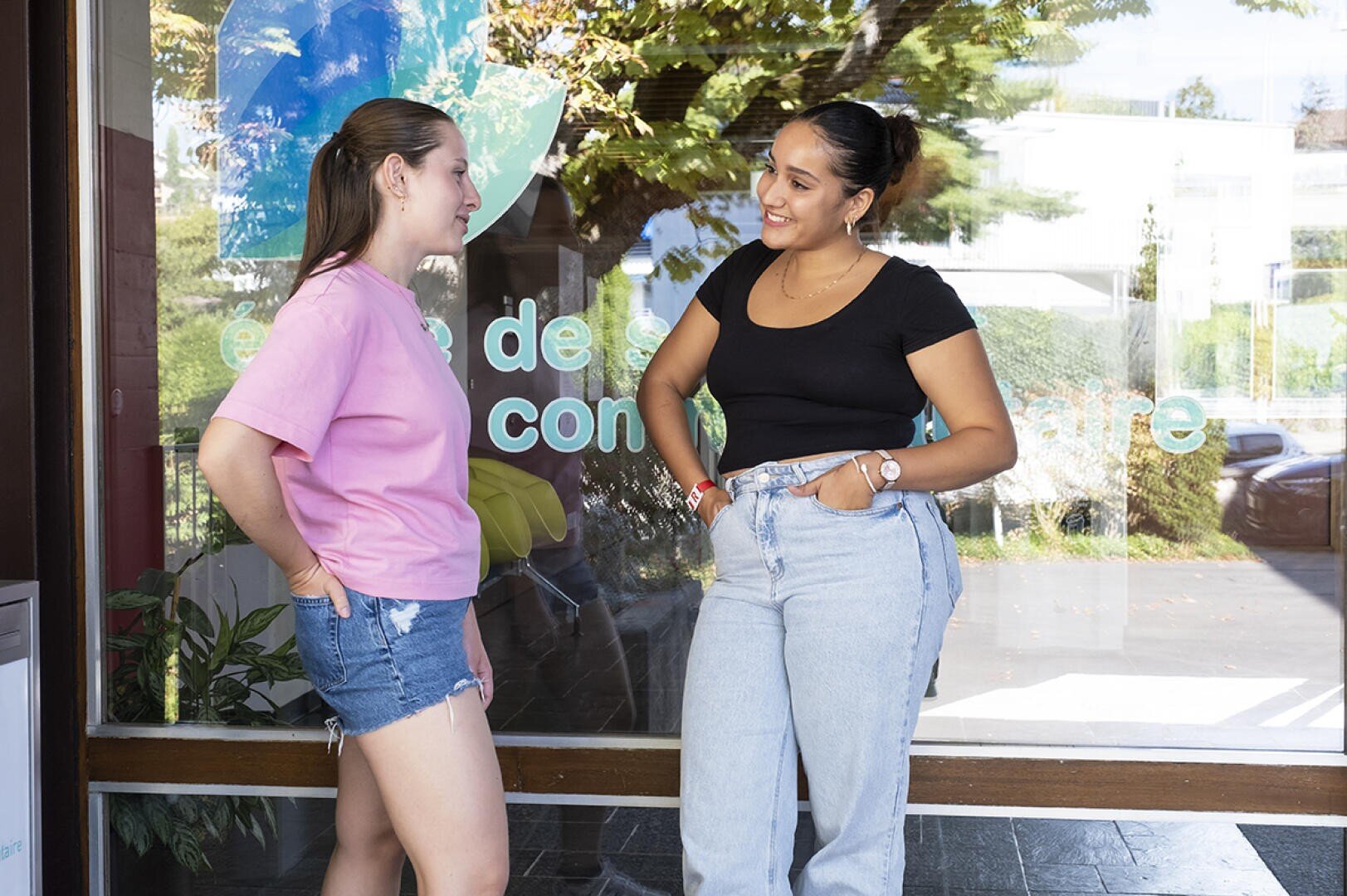 Deux jeunes femmes se tiennent dehors devant une baie vitrée, souriantes et discutant. L'une porte un t-shirt rose et un short en jean, l'autre un t-shirt noir et un jean. Des arbres et des reflets sont visibles en arrière-plan.