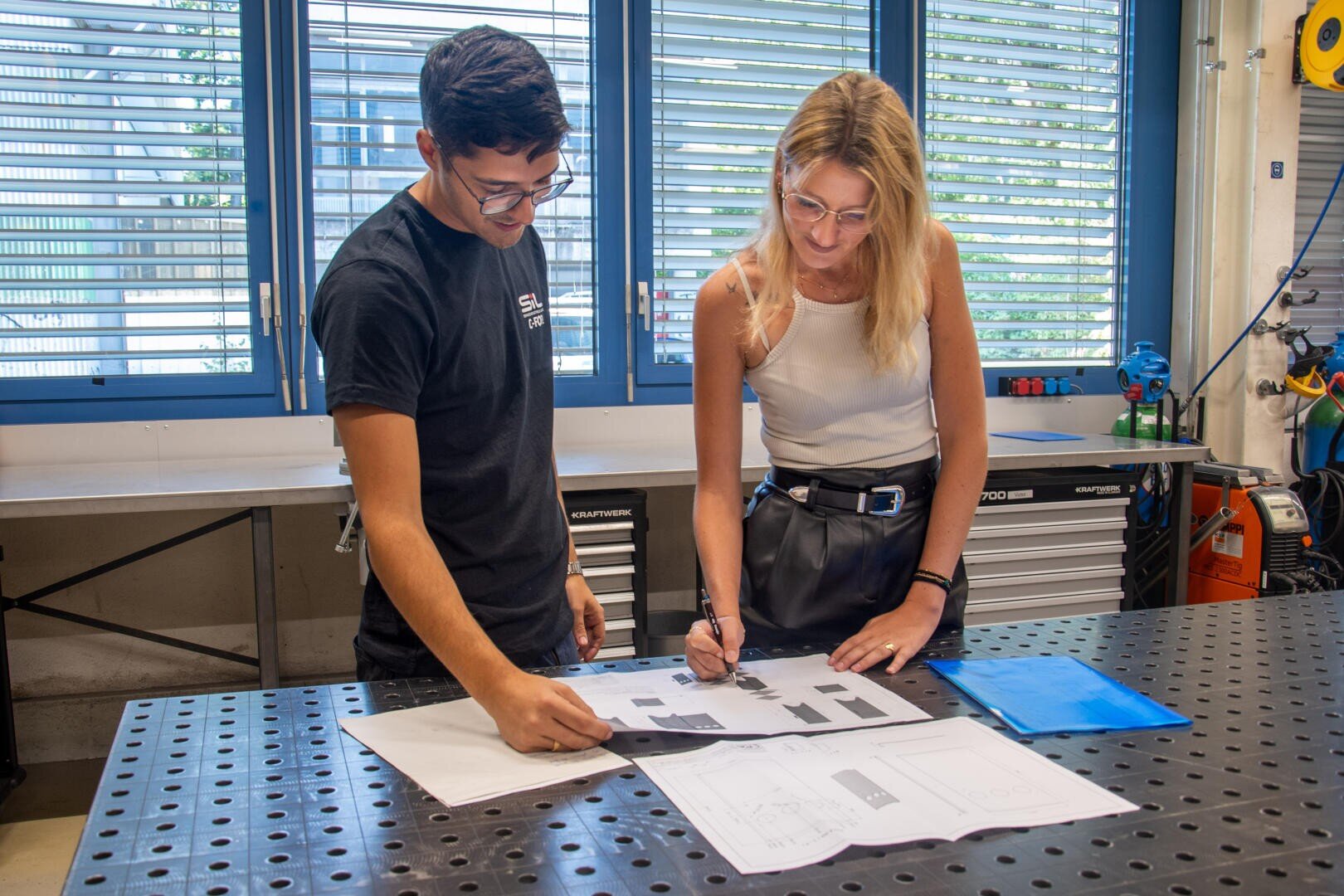 Dans un atelier, deux personnes se tiennent à un établi et examinent des plans d'architecture. L'un pointe du doigt tandis que l'autre écrit. Des outils et du matériel sont visibles à l'arrière-plan et de grandes fenêtres laissent entrer la lumière naturelle.