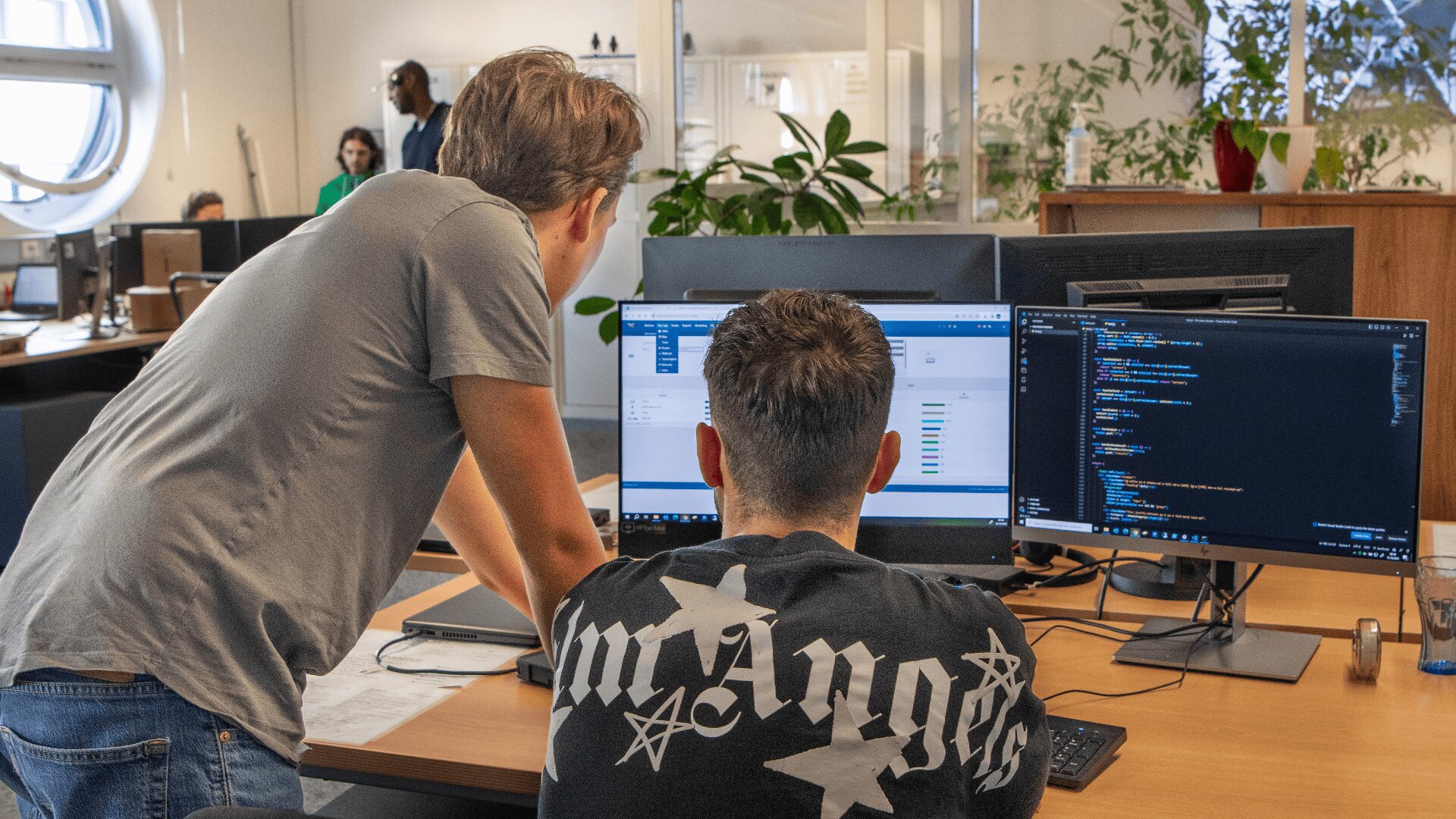 Deux personnes collaborent à un bureau avec deux moniteurs affichant du code et des données dans un espace de bureau avec plusieurs autres personnes travaillant en arrière-plan.