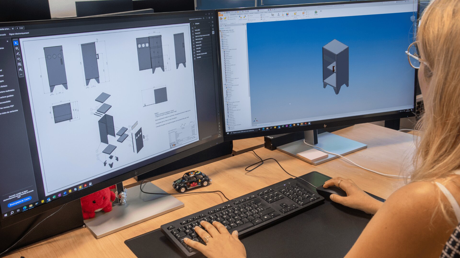 Une personne travaille à un bureau équipé de deux moniteurs ; un écran affiche des diagrammes et des dessins techniques, tandis que l'autre montre un modèle en 3D d'une armoire. Le bureau est équipé d'un clavier, d'une souris et d'une petite voiture.