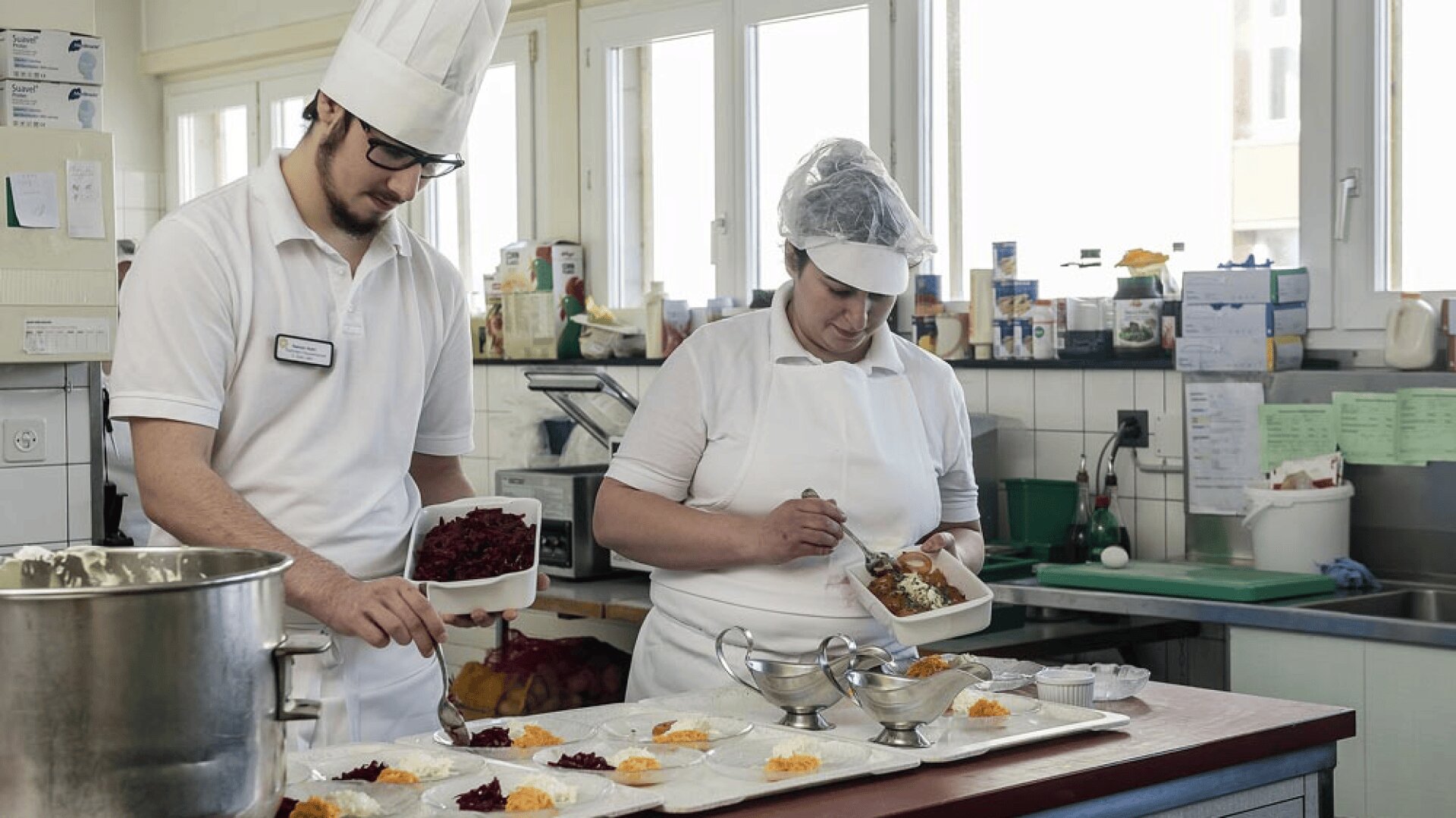 Deux cuisiniers en uniforme blanc et coiffés d'un chapeau préparent et disposent les aliments dans une cuisine claire et propre. L'un d'eux sert les aliments dans des assiettes tandis que l'autre ajoute de la salade à partir d'un récipient. Divers ustensiles de cuisine et ingrédients sont visibles.