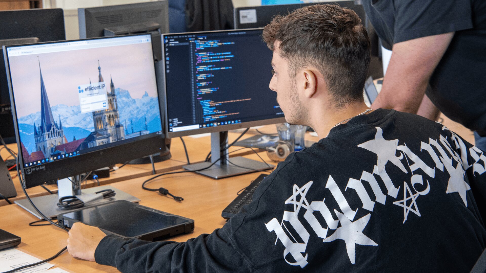 Une personne est assise à un bureau et utilise un ordinateur doté de deux moniteurs, l'un affichant un code et l'autre un écran de connexion. Une autre personne se tient à proximité, partiellement visible, et propose son aide.