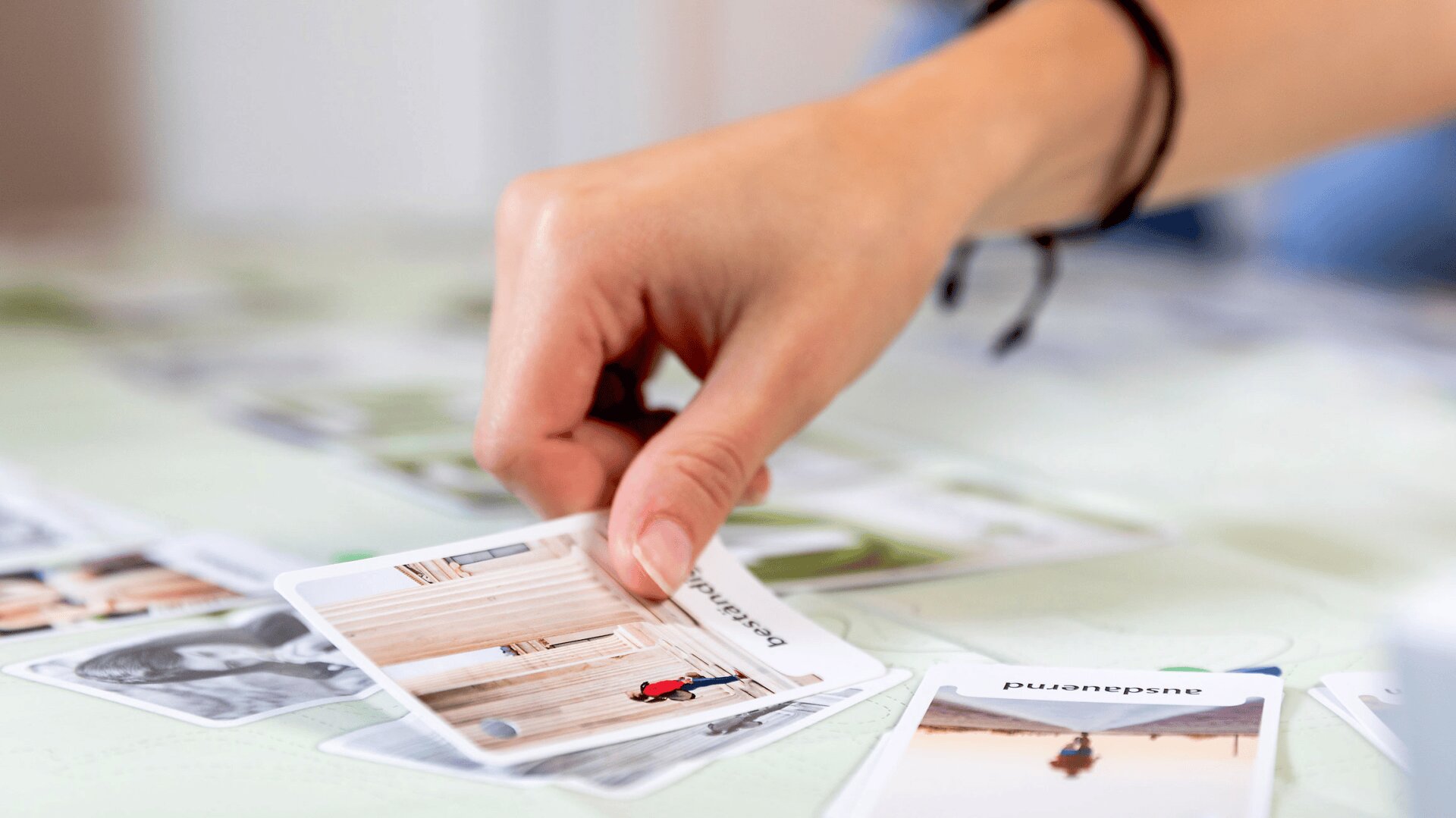 Gros plan d'une main qui prend une carte avec une photo et un texte sur une table parsemée de diverses cartes, chacune affichant des images et des mots différents. L'accent est mis sur la main et les cartes, avec un arrière-plan doux.