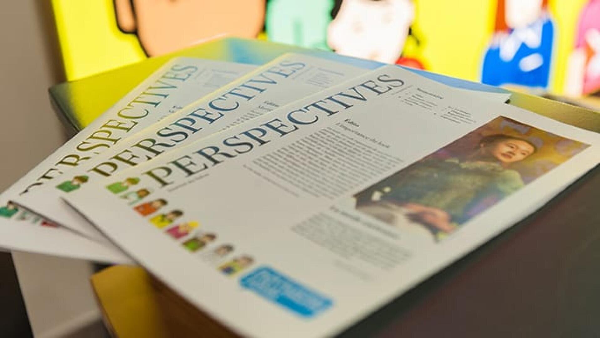 Gros plan sur trois exemplaires d'un magazine intitulé Perspectives, posés sur une table, avec en couverture la photo d'une femme. À l'arrière-plan, on aperçoit partiellement des œuvres d'art colorées.