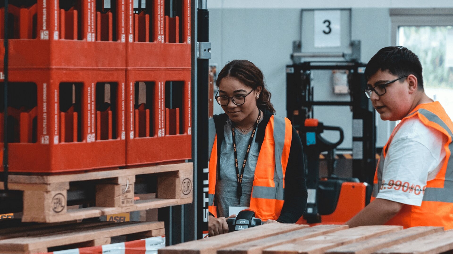 Deux personnes portant des gilets de sécurité orange travaillent ensemble dans un entrepôt, debout à côté de palettes en bois et de caisses rouges, avec des équipements et des rayonnages visibles à l'arrière-plan.