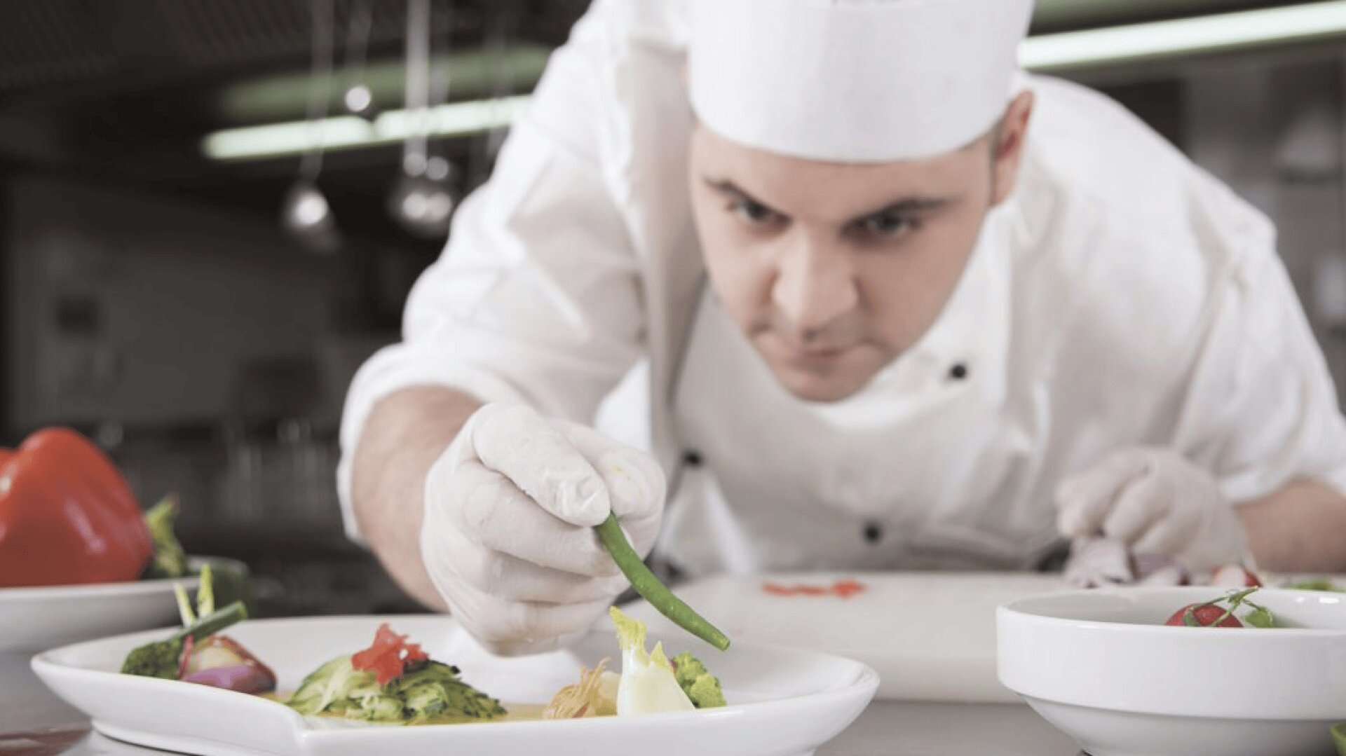 Un chef en uniforme et toque blancs prépare soigneusement un plat, en plaçant à l'aide de pinces un haricot vert sur une assiette blanche contenant divers légumes colorés. L'arrière-plan de la cuisine est légèrement flou.