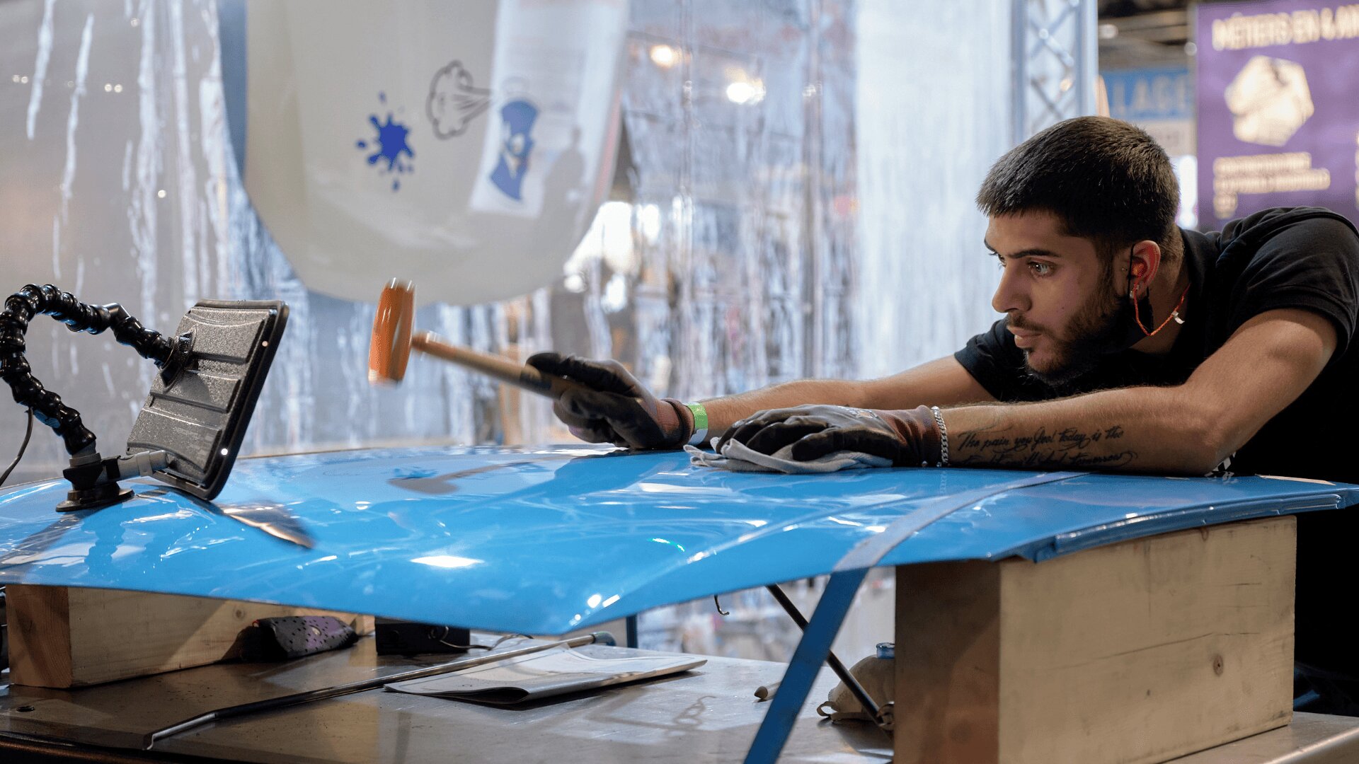 Un homme portant des gants et des protections auditives utilise un maillet pour façonner une grande feuille de métal bleu sur un établi dans un environnement industriel. Il semble concentré et se penche vers l'avant tout en travaillant.