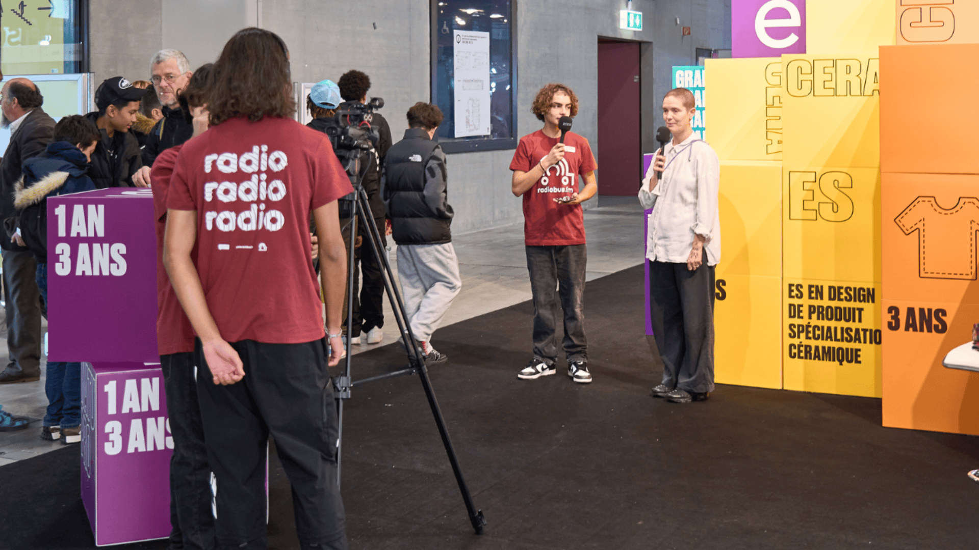 Deux personnes se tiennent sur une scène avec des microphones et s'adressent à un public. Une personne munie d'une caméra et d'autres personnes portant des chemises "radio" les filment. Des panneaux d'affichage colorés avec des textes sur le design et la céramique se trouvent à l'arrière-plan.