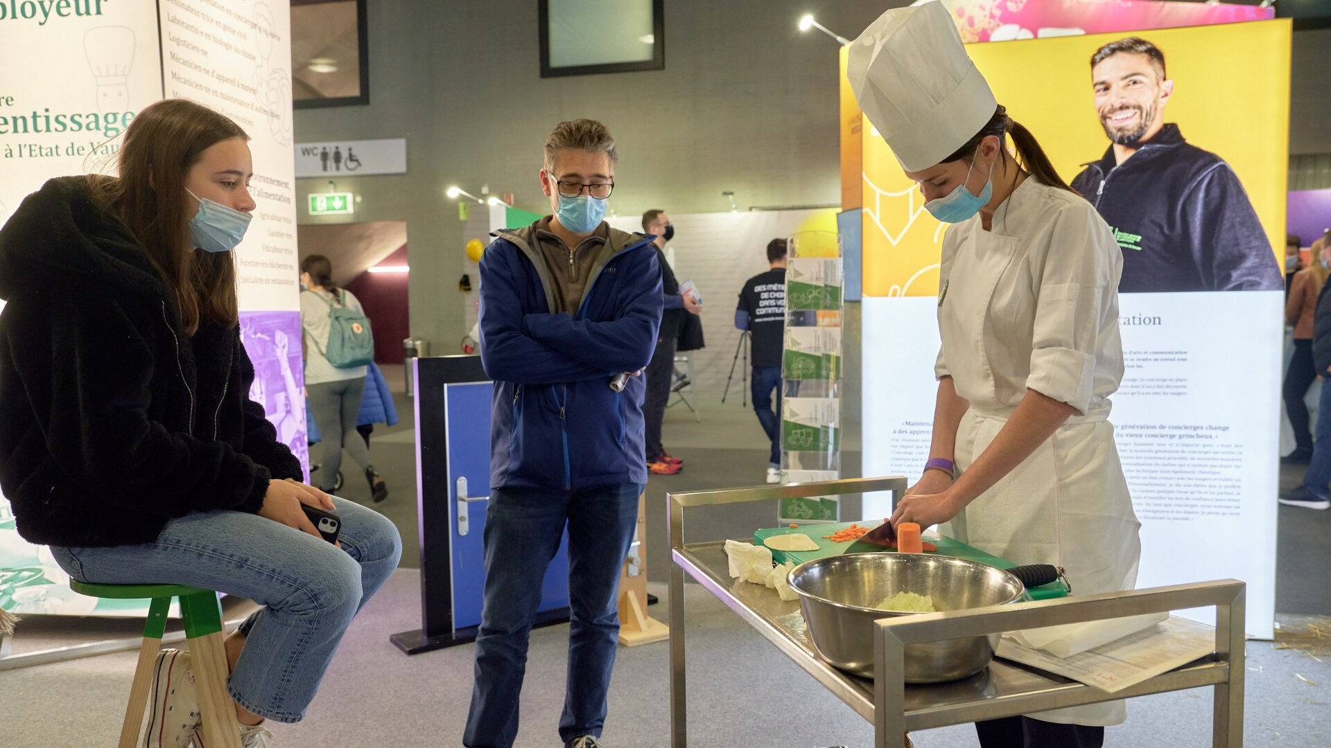 Un chef cuisinier portant un uniforme et un chapeau blancs démontre une technique de cuisine à une table tandis que deux personnes, toutes masquées, regardent attentivement lors d'un événement ou d'une exposition en intérieur. Des ustensiles de cuisine et un bol à mélanger sont posés sur la table.