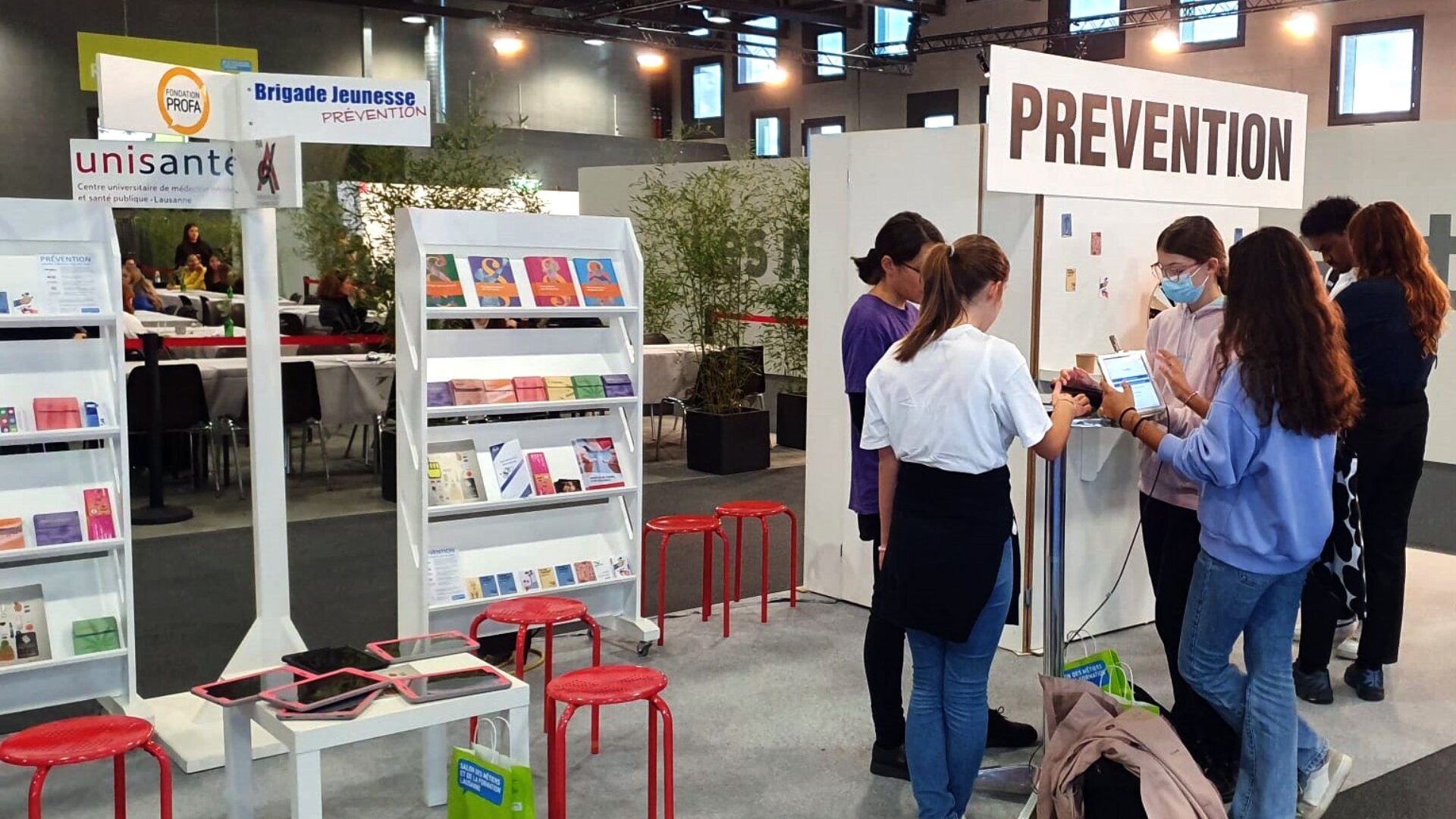 Plusieurs jeunes se tiennent à un stand "PREVENTION" avec des brochures et des tablettes numériques. À proximité, des étagères présentent des brochures sous un panneau "unisante". Des tabourets rouges sont disséminés dans un espace événementiel intérieur.