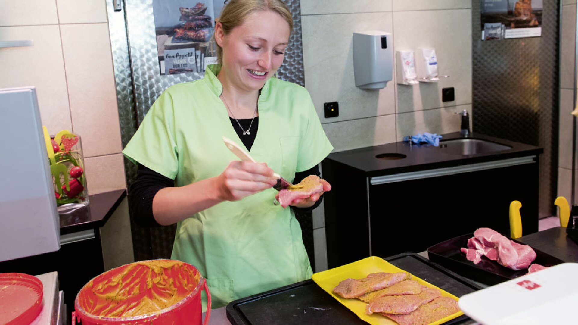 Une femme en uniforme vert clair étale de la marinade sur des tranches de viande crue au comptoir d'une boucherie, tout en souriant. Divers plateaux de viande sont posés sur le comptoir devant elle.