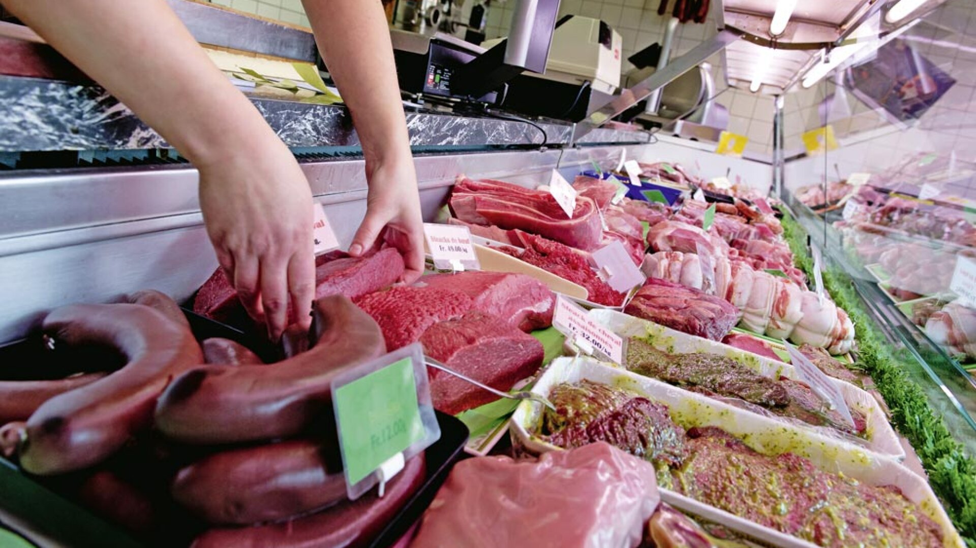 Dans une boucherie ou un comptoir à viande, une personne dispose différents morceaux de viande crue, des saucisses et des steaks dans une vitrine en verre. Des panneaux avec des étiquettes et des prix sont visibles parmi les viandes.
