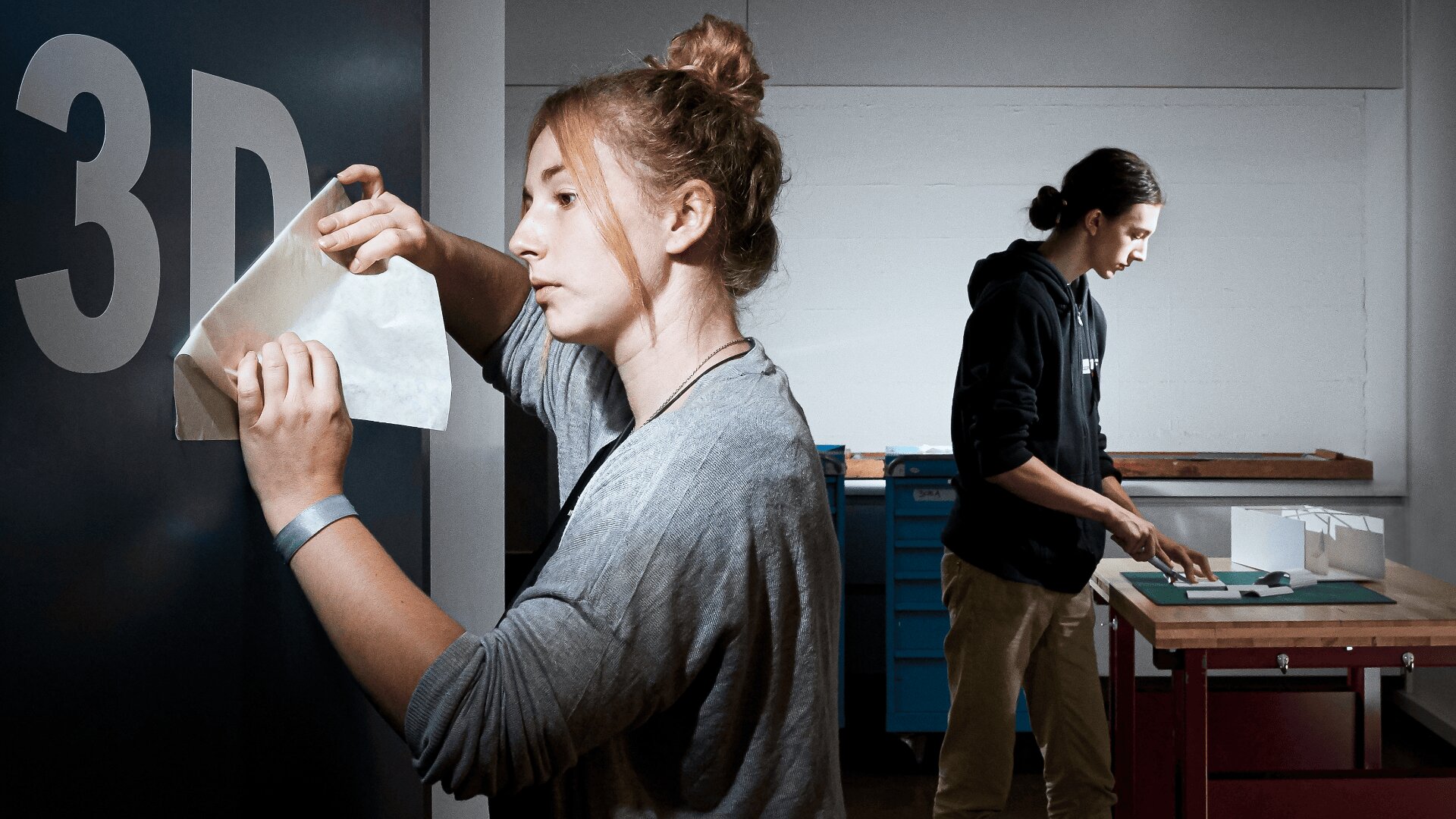 Une jeune femme applique un autocollant en vinyle indiquant "3D" sur un mur, tandis qu'un jeune homme travaille sur un modèle à une table à l'arrière-plan, dans un atelier bien éclairé.