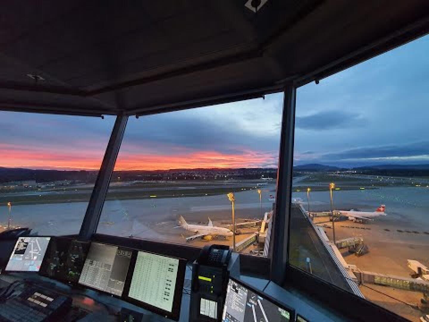 Vue d'une tour de contrôle d'aéroport au coucher du soleil, surplombant des avions stationnés à des portes d'embarquement sur la piste, avec des écrans d'ordinateur au premier plan et un ciel coloré à l'arrière-plan.