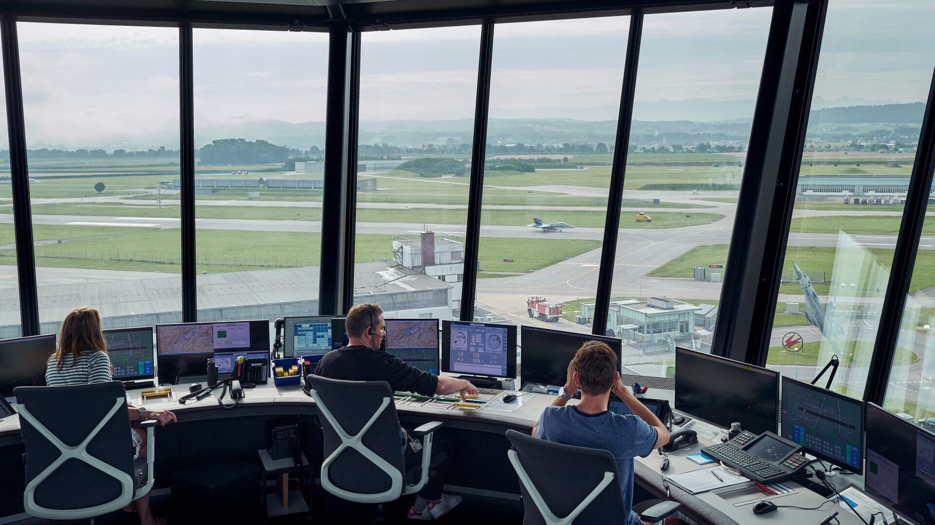 Trois contrôleurs aériens sont assis à des bureaux équipés de plusieurs écrans dans une tour de contrôle, surveillant les avions sur les pistes et les voies de circulation à travers de grandes fenêtres offrant une vue étendue sur l'aéroport et le paysage lointain.