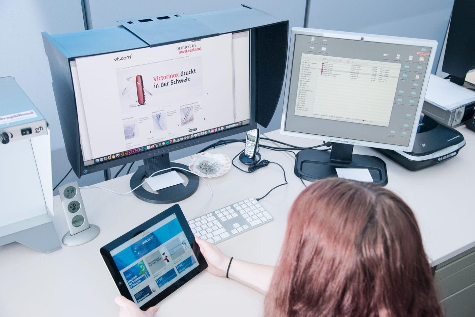 Une personne aux longs cheveux bruns est assise à un bureau et regarde une tablette tandis que deux moniteurs d'ordinateur affichent différents écrans. Le bureau comporte également un clavier, une souris, un téléphone et quelques articles de bureau.