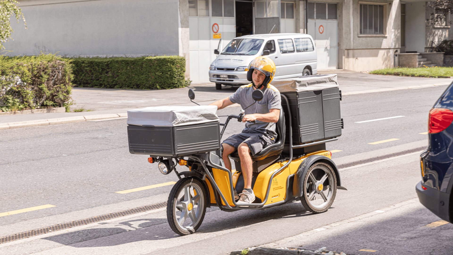Une personne portant un casque jaune conduit un scooter de livraison jaune à trois roues avec de grandes boîtes de rangement à l'avant et à l'arrière, dans une rue d'une zone urbaine.