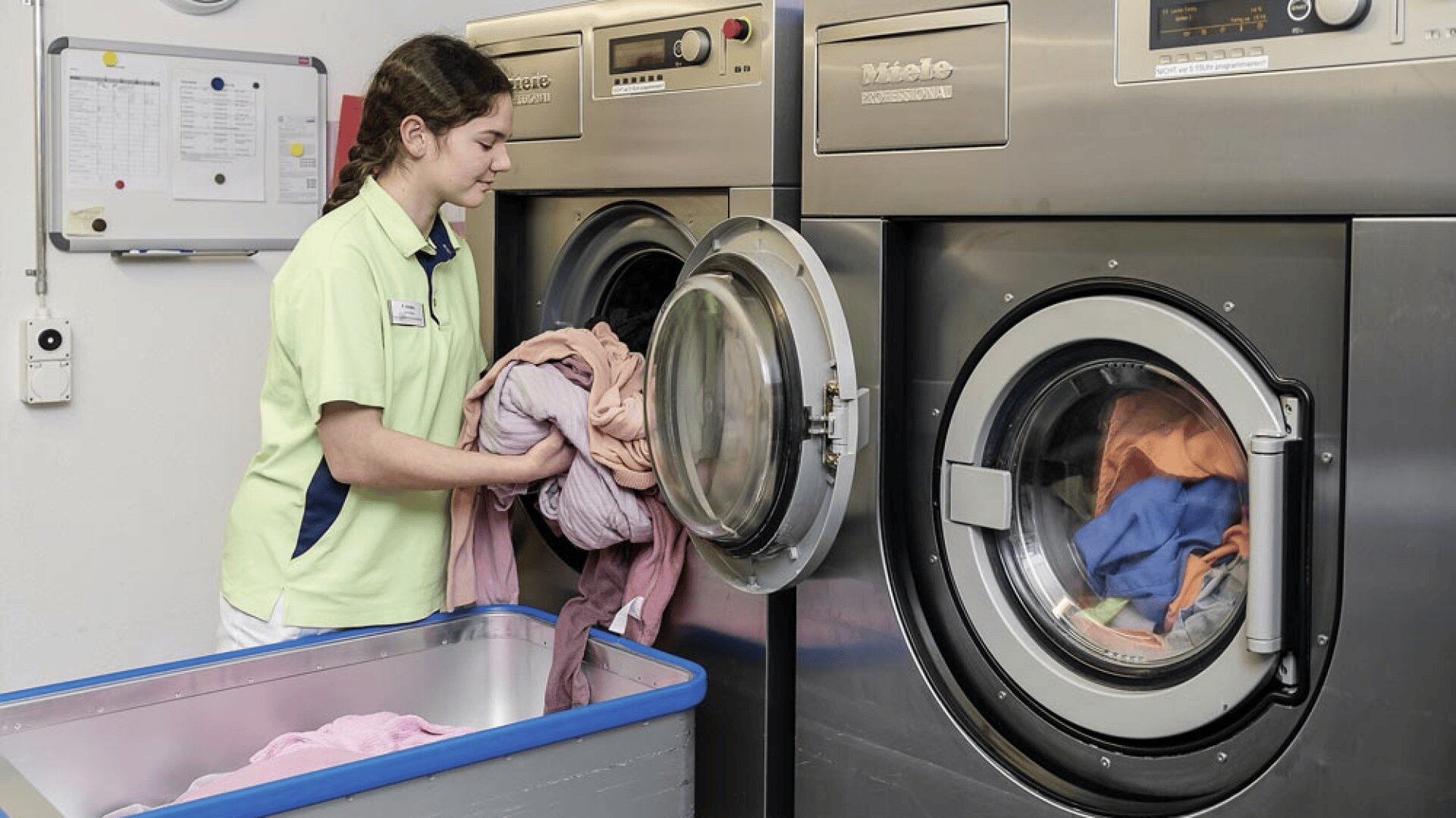 Une femme portant un uniforme vert clair charge du linge coloré d'un chariot bleu dans une machine à laver à chargement frontal dans une buanderie. Une autre machine à laver avec des vêtements à l'intérieur se trouve à côté d'elle.