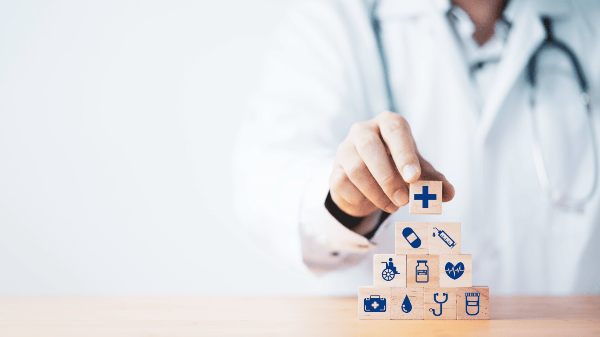 Un médecin en blouse blanche empile des blocs de bois avec des icônes de soins de santé, formant une pyramide. Le bloc supérieur comporte une croix médicale bleue, qui symbolise les soins de santé et les services médicaux.