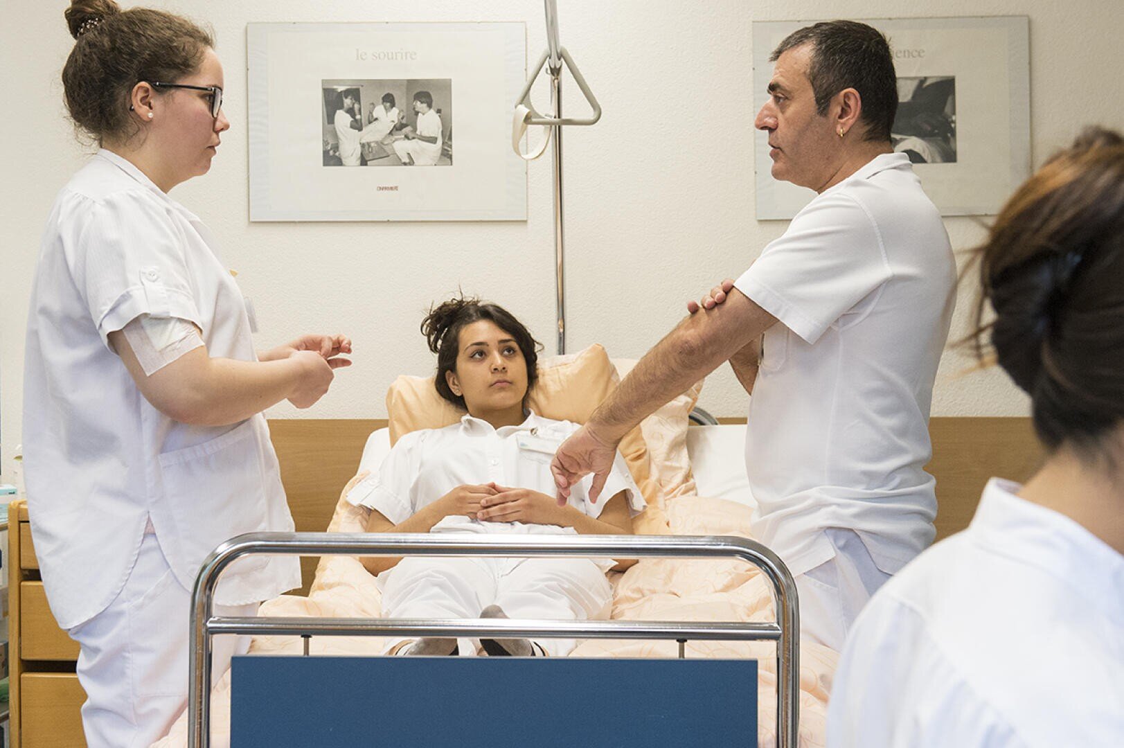 Trois professionnels de la santé, deux debout et un assis, discutent au chevet d'un patient allongé dans un lit d'hôpital, dans une salle d'hôpital lumineuse avec des affiches médicales au mur.