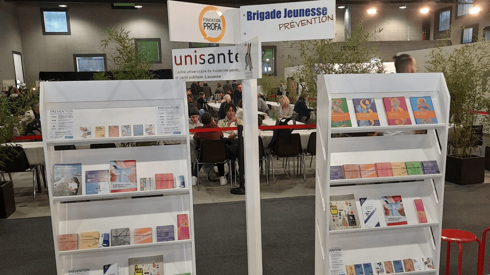 Un présentoir avec des brochures et des livrets sur la santé et la prévention, étiqueté Brigade Jeunesse PRÉVENTION, dans un espace événementiel intérieur très fréquenté, avec des personnes assises à des tables à l'arrière-plan.