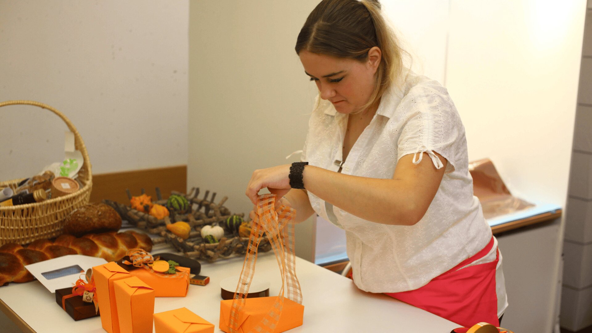 Une femme vêtue d'un chemisier blanc et d'une jupe rose noue un ruban autour d'un paquet cadeau orange posé sur une table, avec d'autres paquets orange et des articles de boulangerie assortis à l'arrière-plan.