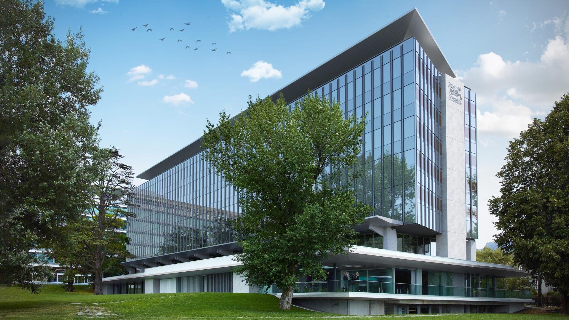 Un immeuble de bureaux moderne en verre se dresse derrière des arbres verts et une pelouse, sous un ciel bleu parsemé de nuages et d'oiseaux en vol.