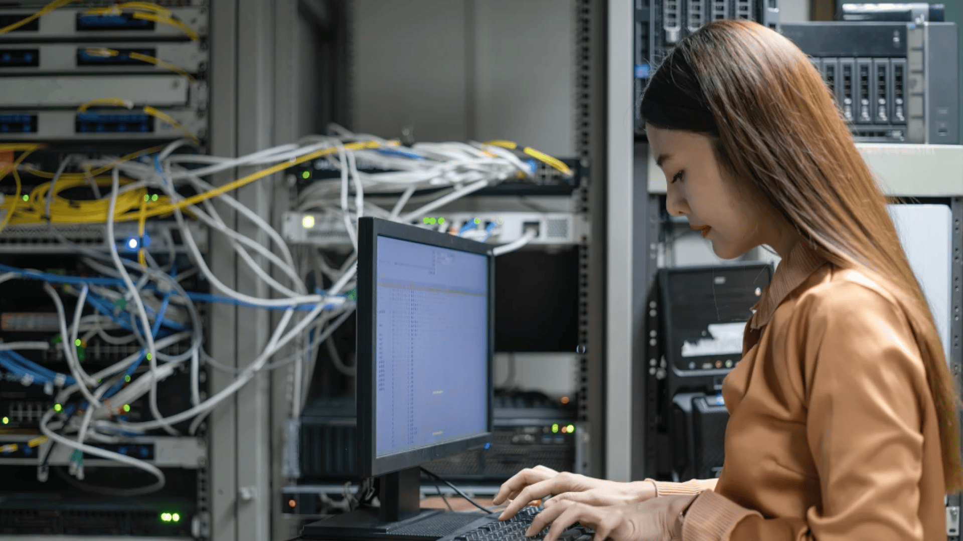 Une femme est assise à un bureau et tape sur un ordinateur dans une salle de serveurs, entourée de câbles de réseau et d'équipements de serveurs.