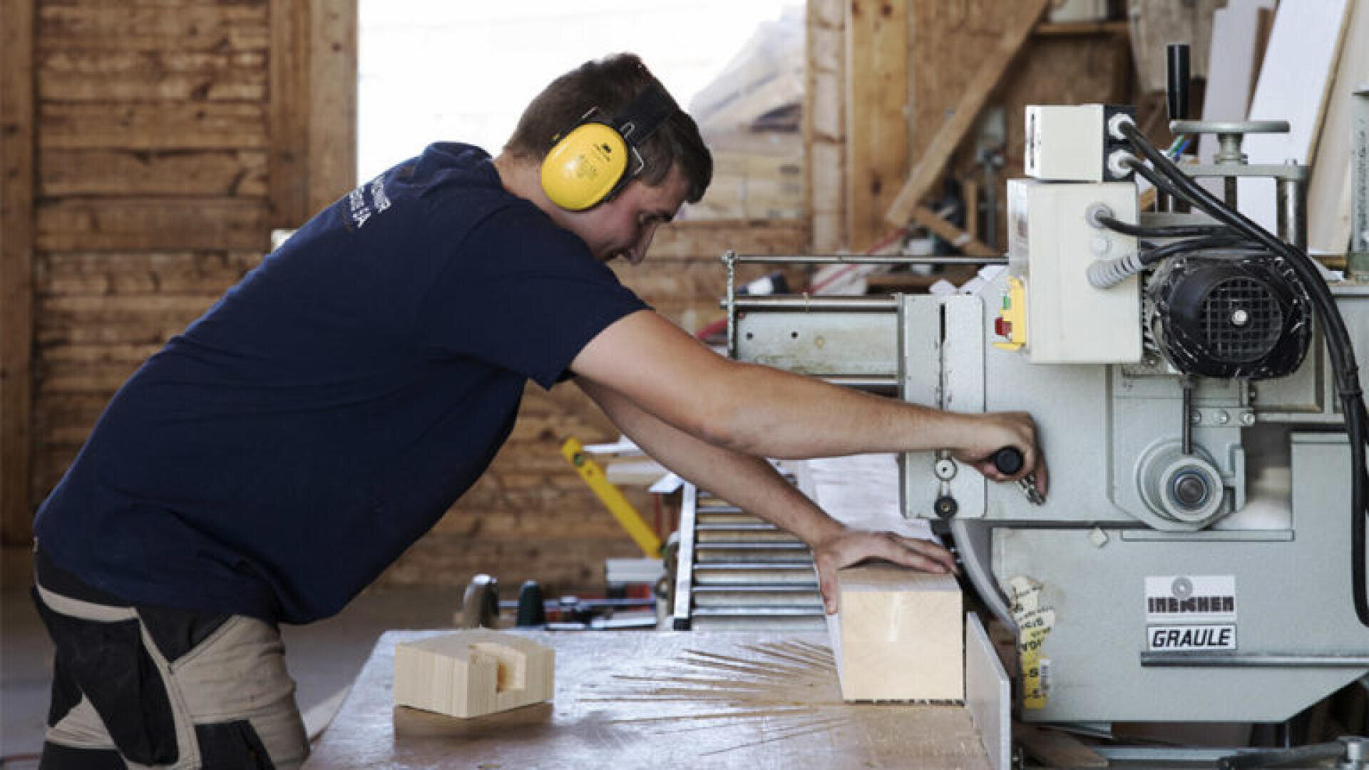 Un homme portant une protection auditive jaune fait fonctionner une grande machine à travailler le bois dans un atelier, en ajustant un bloc de bois sur la surface de travail.