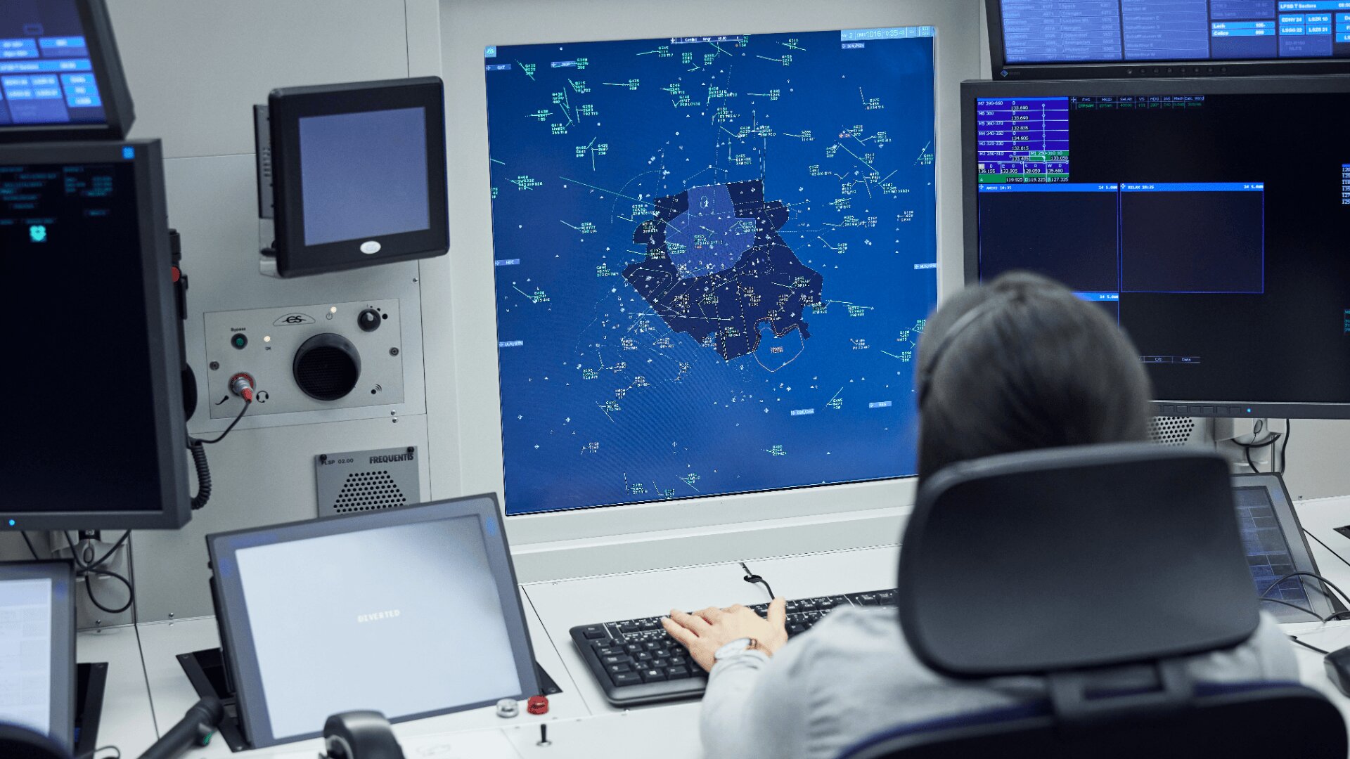 Une personne est assise à un bureau devant un grand écran radar affichant des données de contrôle du trafic aérien, entourée de plusieurs moniteurs et d'équipements techniques.