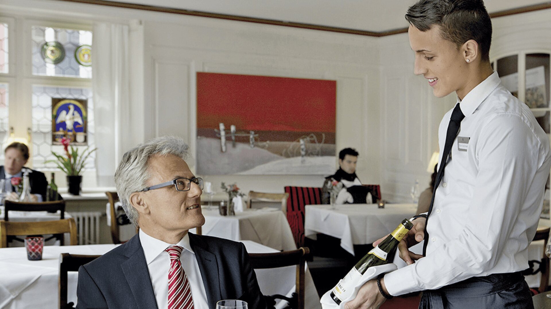 Un serveur souriant en chemise blanche et cravate noire présente une bouteille de vin à un homme plus âgé en costume et lunettes, assis à une table dans un élégant restaurant aux nappes blanches et aux accents rouges.