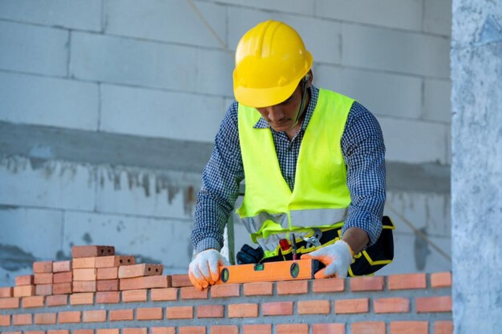Un ouvrier du bâtiment portant un casque jaune et un gilet de sécurité utilise un niveau à bulle pour vérifier l'alignement des briques sur un mur de briques partiellement construit sur un chantier de construction.