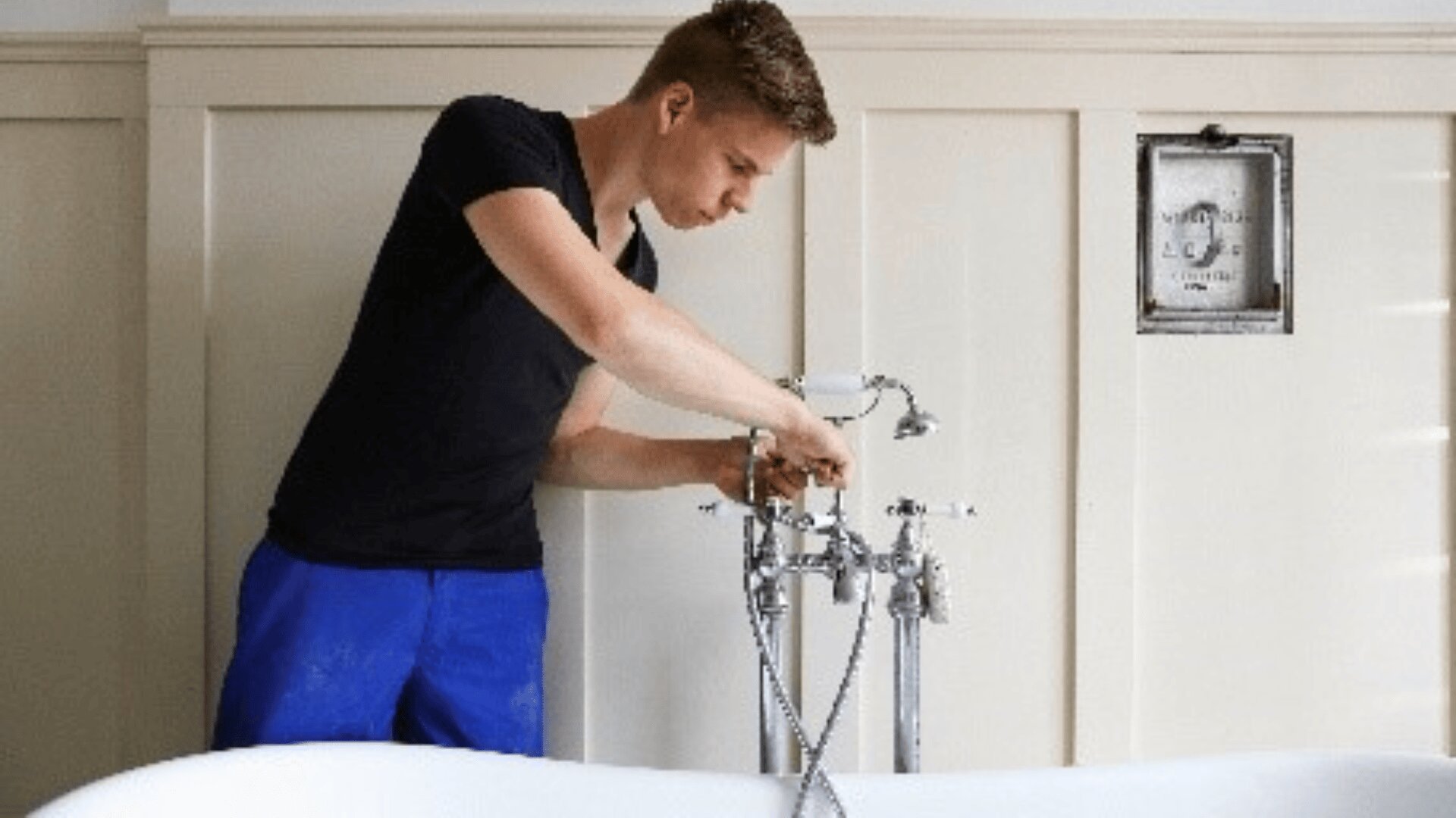 Un jeune homme vêtu d'un tee-shirt noir et d'un pantalon bleu ajuste le robinet d'une baignoire dans une salle de bains lumineuse aux murs blancs.