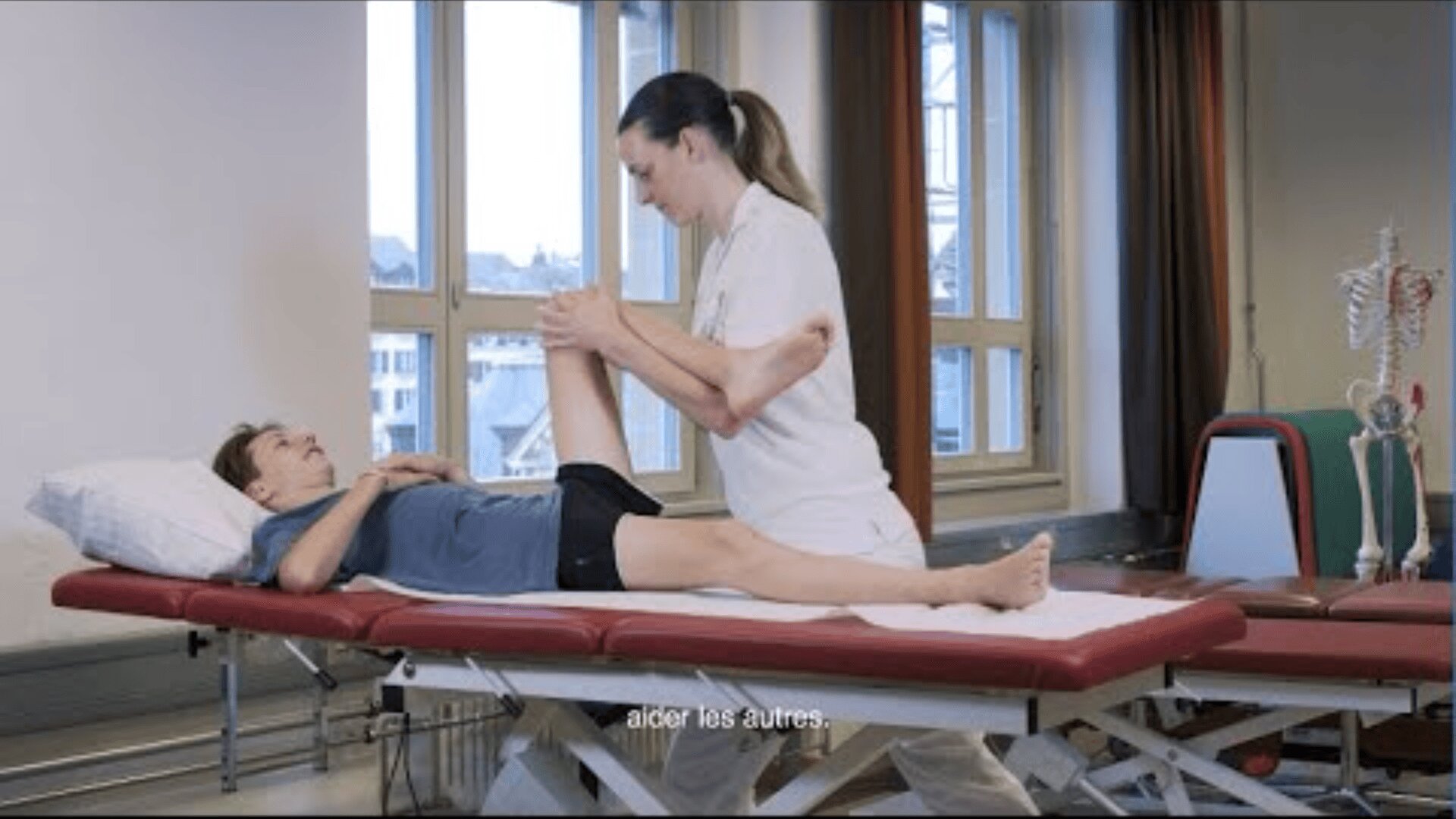 Un kinésithérapeute en uniforme blanc aide un patient allongé sur une table de traitement à étirer sa jambe. La pièce est dotée de grandes fenêtres, d'une maquette de squelette et d'équipements médicaux. Des sous-titres en français sont visibles en bas de l'image.
