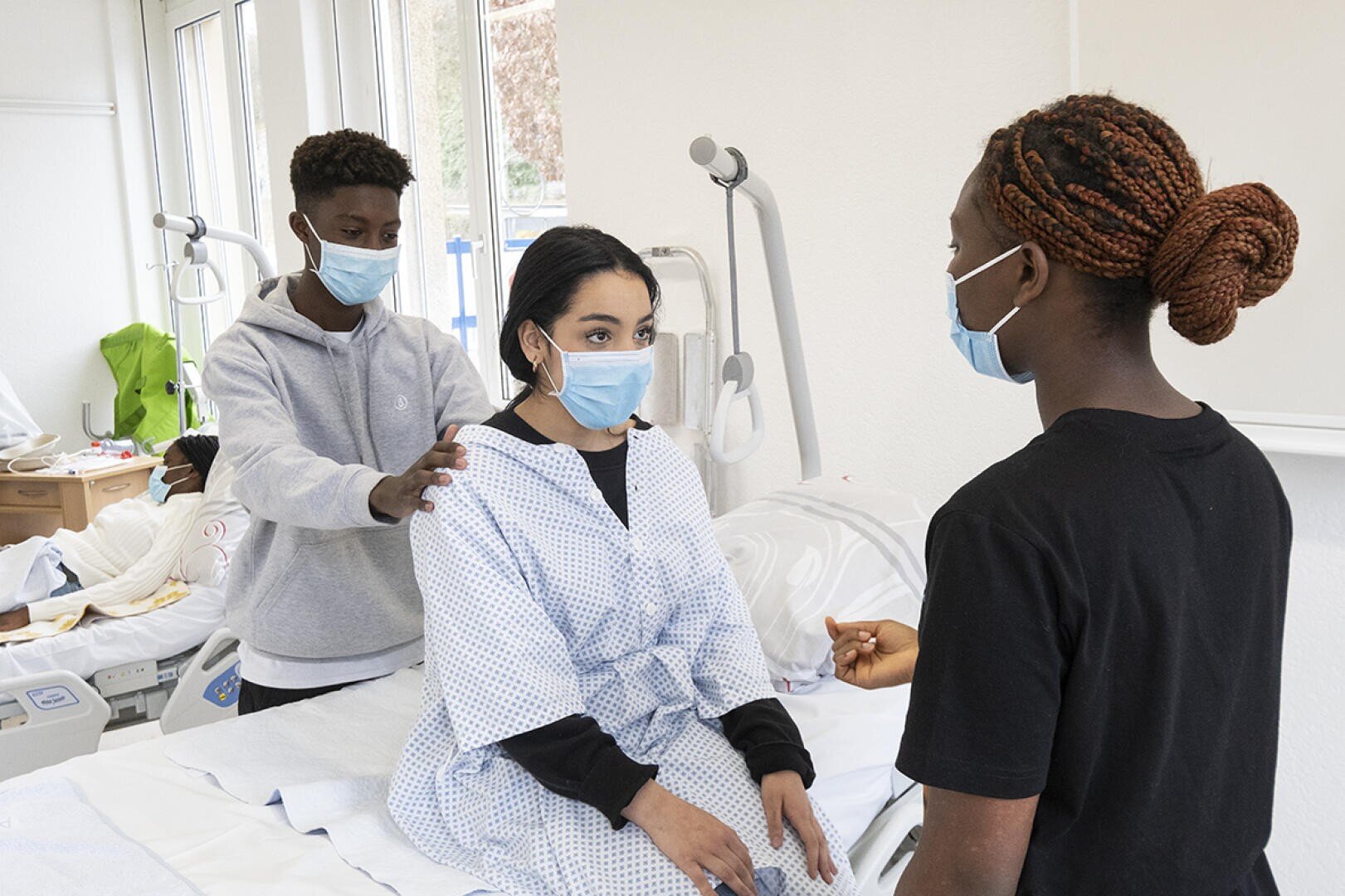Trois personnes portant des masques se trouvent dans une chambre d'hôpital. Une personne vêtue d'une blouse est assise sur un lit, tandis qu'une autre se tient derrière, leur touchant les épaules. Une troisième personne se tient face à eux, tenant un objet, comme pour expliquer ou donner des instructions.
