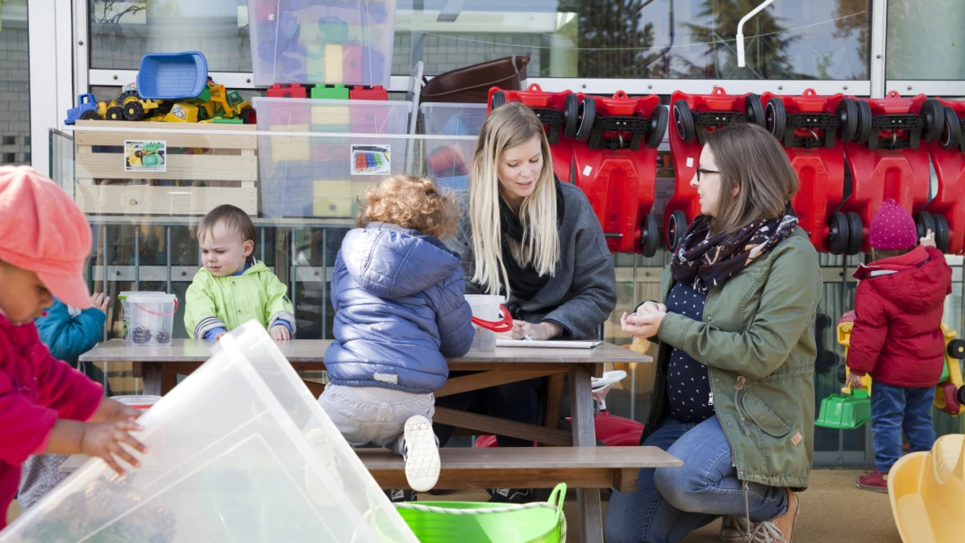 Deux femmes sont assises à une table de pique-nique en plein air et de jeunes enfants jouent autour d'elles. Des jouets colorés et des petites voitures sont visibles à l'arrière-plan. Un enfant en bas âge est assis à la table, et un autre enfant joue à proximité.