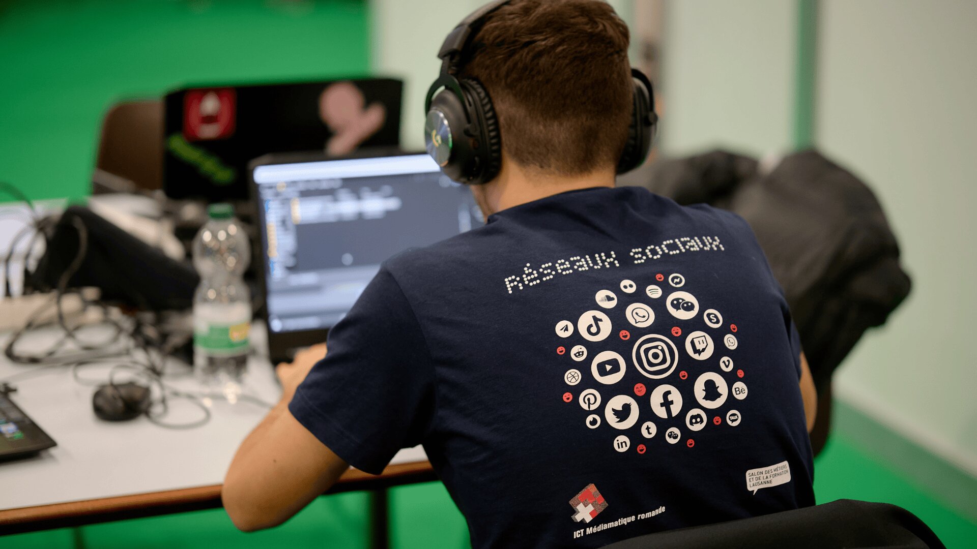 Une personne portant des écouteurs et un t-shirt Réseaux Sociaux avec des icônes de médias sociaux au dos est assise à un bureau, travaillant sur un ordinateur portable. Une bouteille d'eau et un ordinateur portable avec des autocollants sont également visibles.