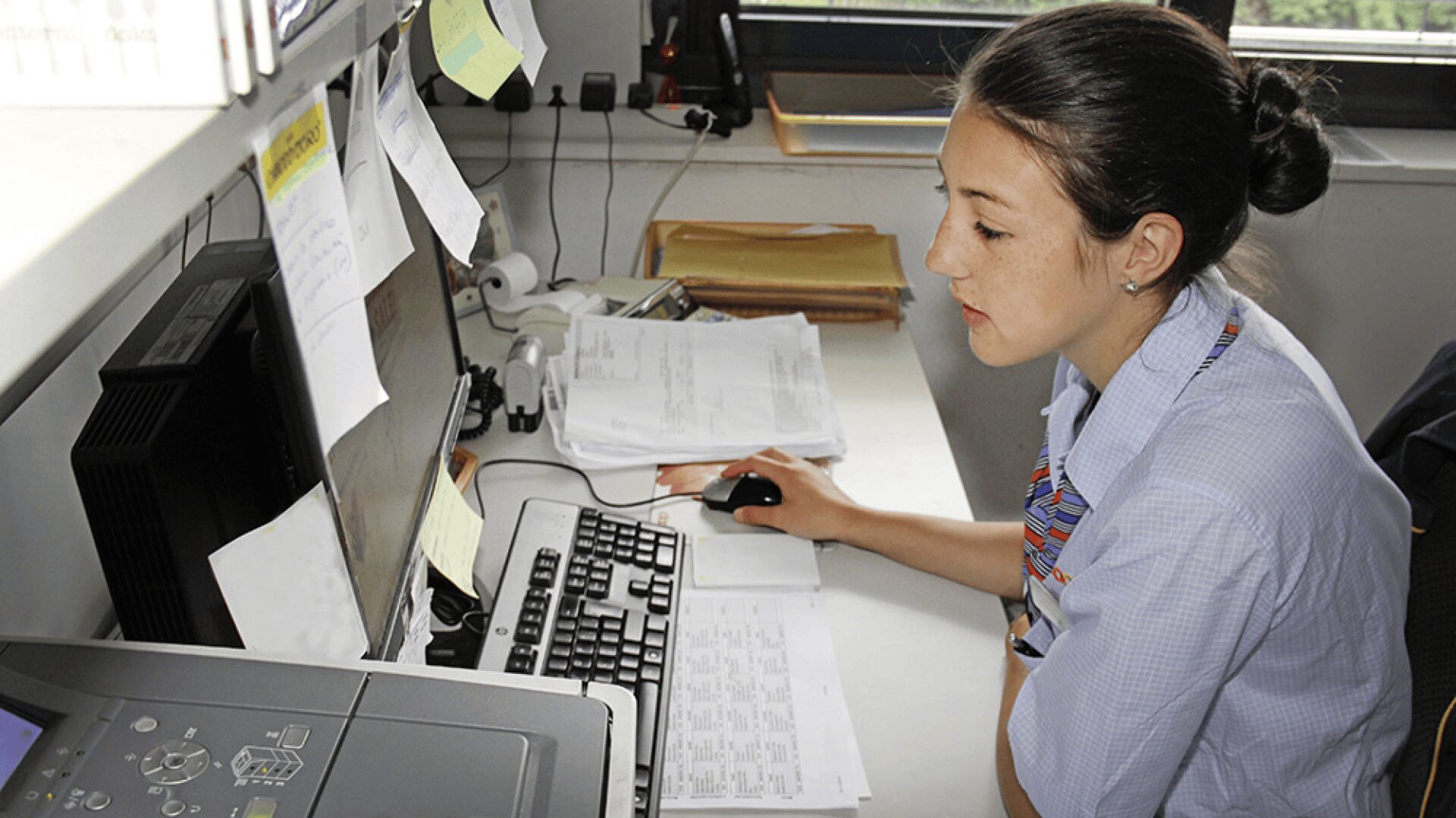 Une femme en uniforme de bureau est assise à un bureau et utilise un ordinateur. Des papiers, des notes autocollantes et des documents entourent son espace de travail, et elle semble concentrée sur l'écran.
