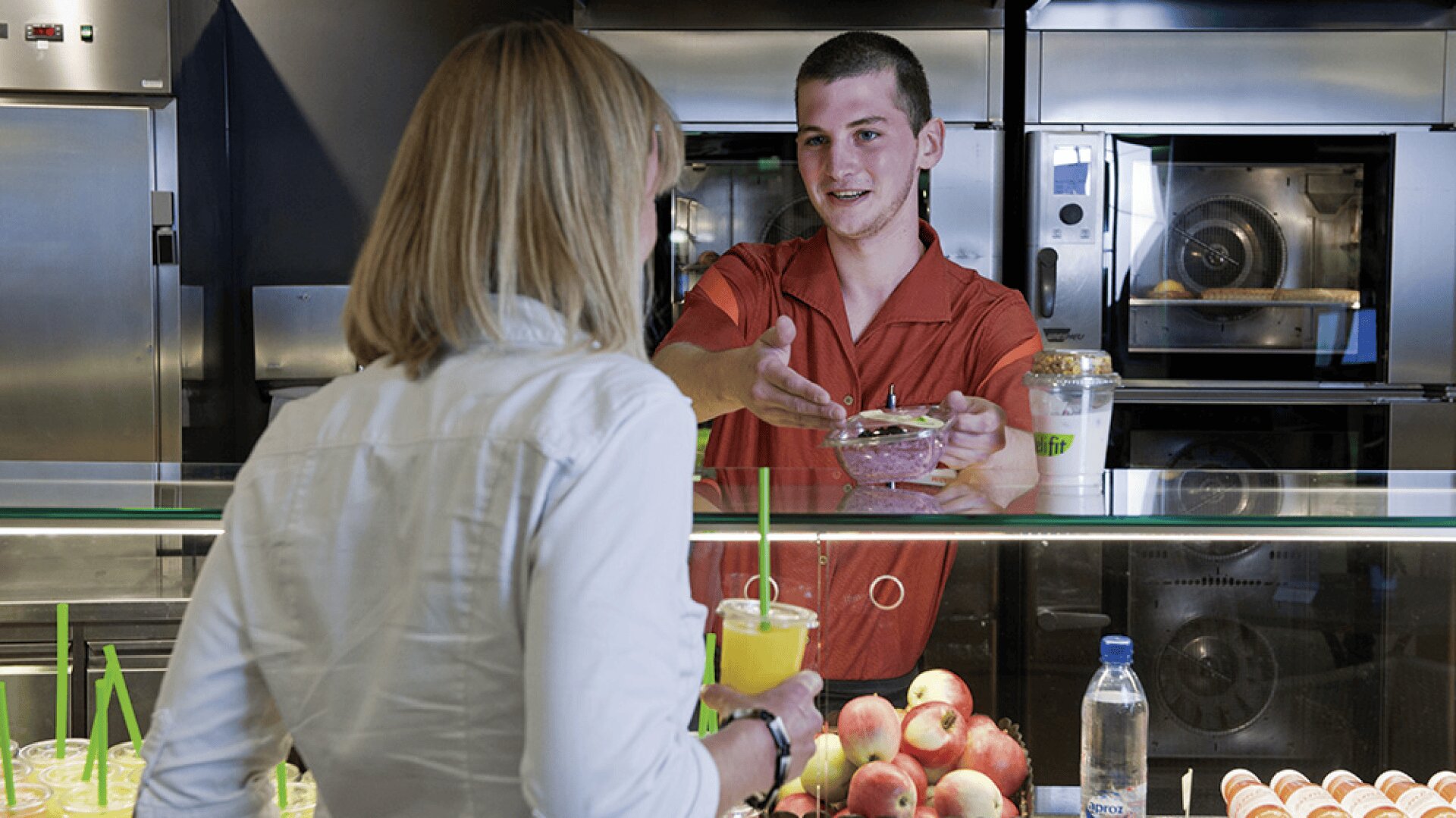 Un jeune homme derrière un comptoir tend un récipient de nourriture à une femme aux cheveux blonds qui tient une boisson. Des fruits frais, des smoothies et des bouteilles d'eau sont présentés sur le comptoir d'un magasin d'alimentation moderne.