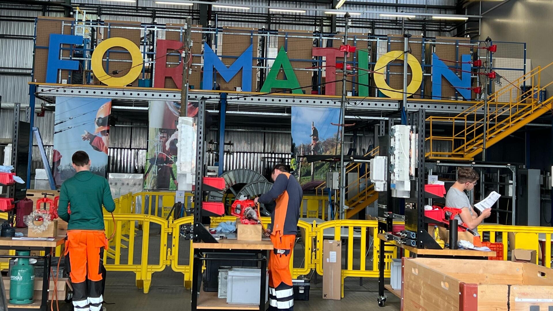 Trois personnes vêtues de pantalons de travail orange et d'équipements de sécurité travaillent à des postes distincts sous un grand panneau coloré FORMATION à l'intérieur d'un centre de formation industrielle doté d'outils et d'équipements.