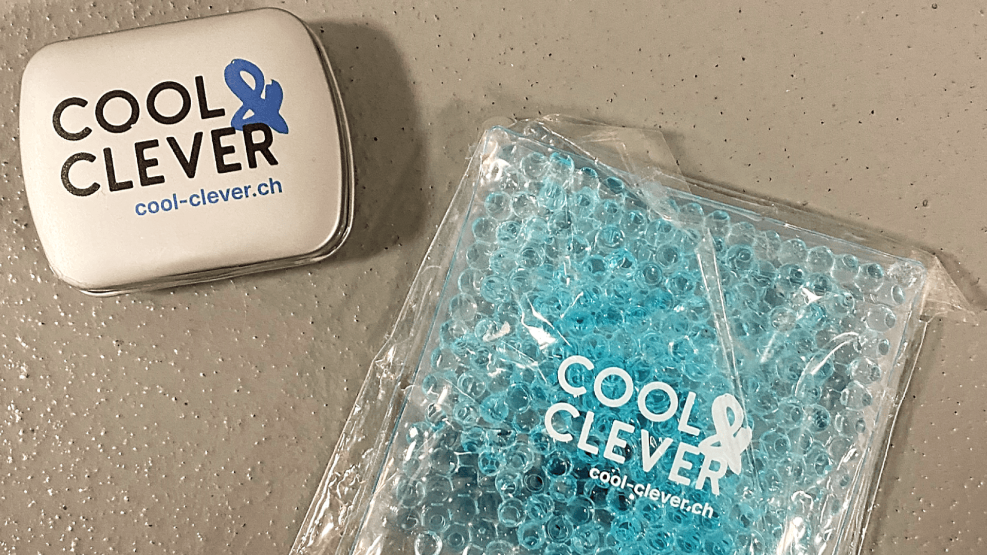 Une petite boîte métallique et un sachet de gel bleu, tous deux étiquetés COOL & CLEVER avec le site web cool-clever.ch, reposent sur une surface grise texturée.